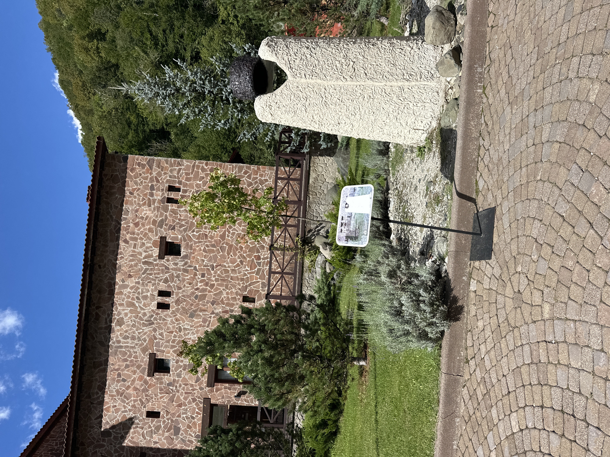 A stone statue stands in a garden in front of a stone building. The statue is a rough-hewn, light-colored figure with a textured surface, topped with a dark, spherical object. A sign with text and an image is placed in front of the statue, on a black metal stand. The building behind the statue is made of rough-hewn stone and has small, dark windows. A wooden fence runs along the base of the building. The garden area includes green grass, bushes, and trees, including a tall, slender evergreen tree. In the background, a densely wooded hillside rises under a blue sky with scattered white clouds. The foreground features a paved walkway made of light-brown cobblestones. The location is identified as Estosadok, Russia.