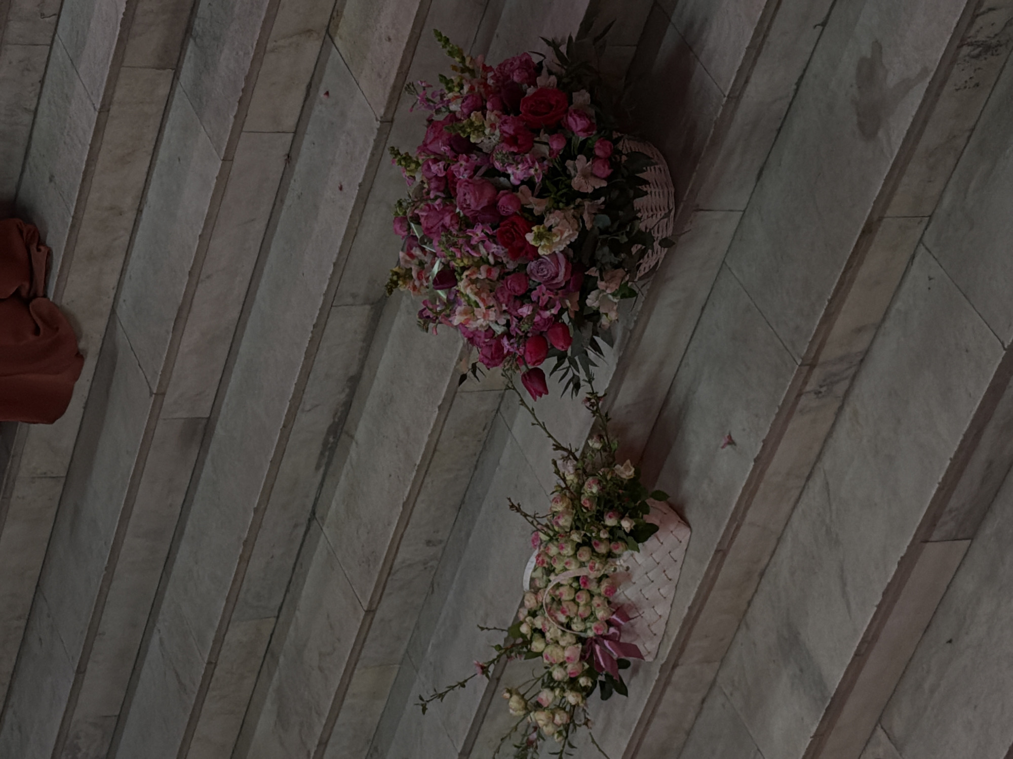 The image depicts a floral arrangement on a gray stone wall. The arrangement consists of two woven baskets, each filled with flowers. The upper basket contains a variety of pink and red roses. The lower basket contains small white flowers and green leaves. The background is a series of gray stone tiles. The location is unknown, and no people, subjects, objects, actions, or interactions are discernible besides the floral arrangement and its immediate surrounding environment. The claim that it is located in Yerevan, Armenia, is not verifiable from the image.