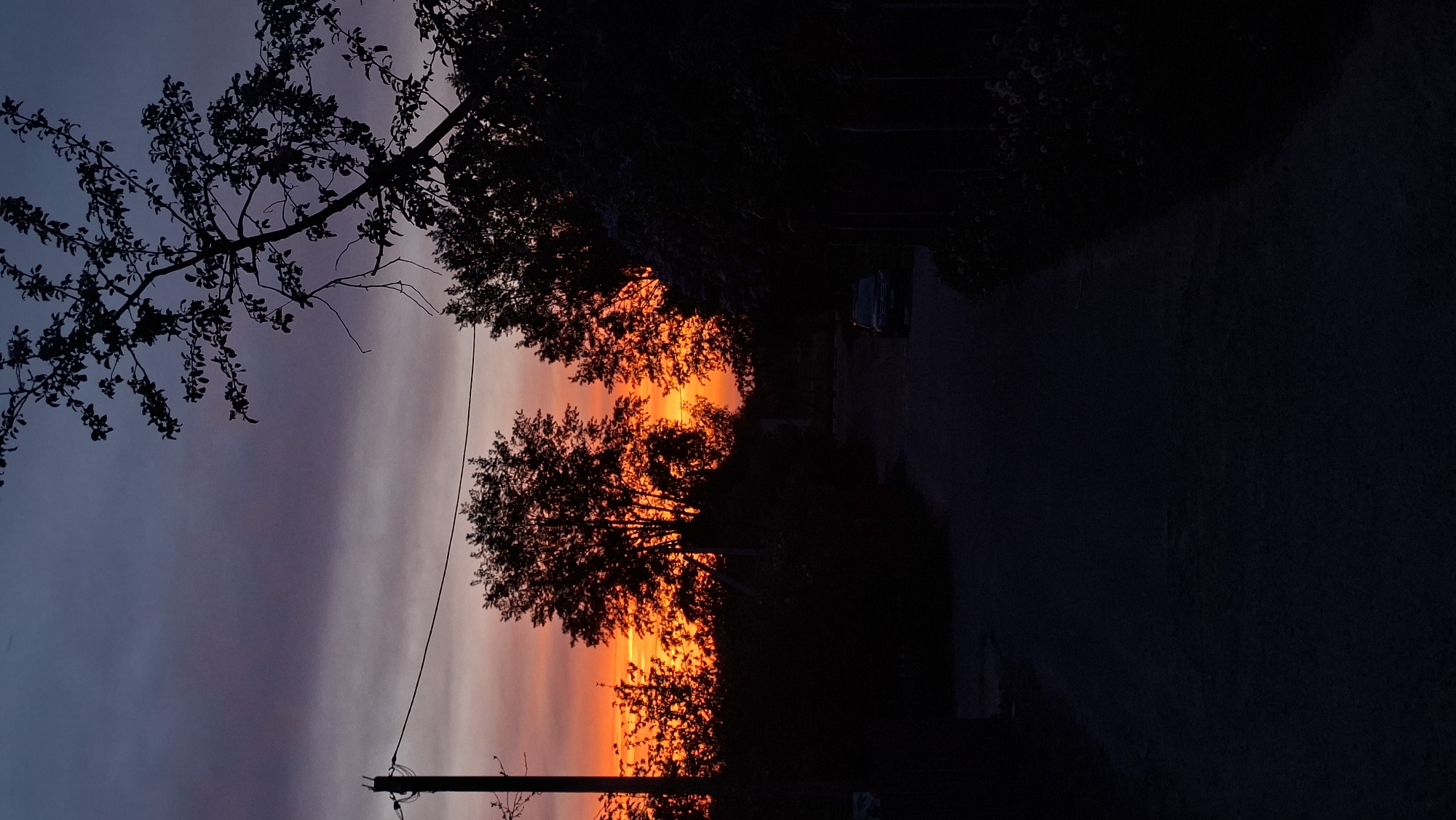 The image depicts a sunset over a road in an unknown location. Trees are silhouetted against a sky with hues of orange, red, purple, and blue. A utility pole with wires is visible on the left side of the image. A car is parked on the side of the road in the distance. The road leads away from the viewer into the darkness.