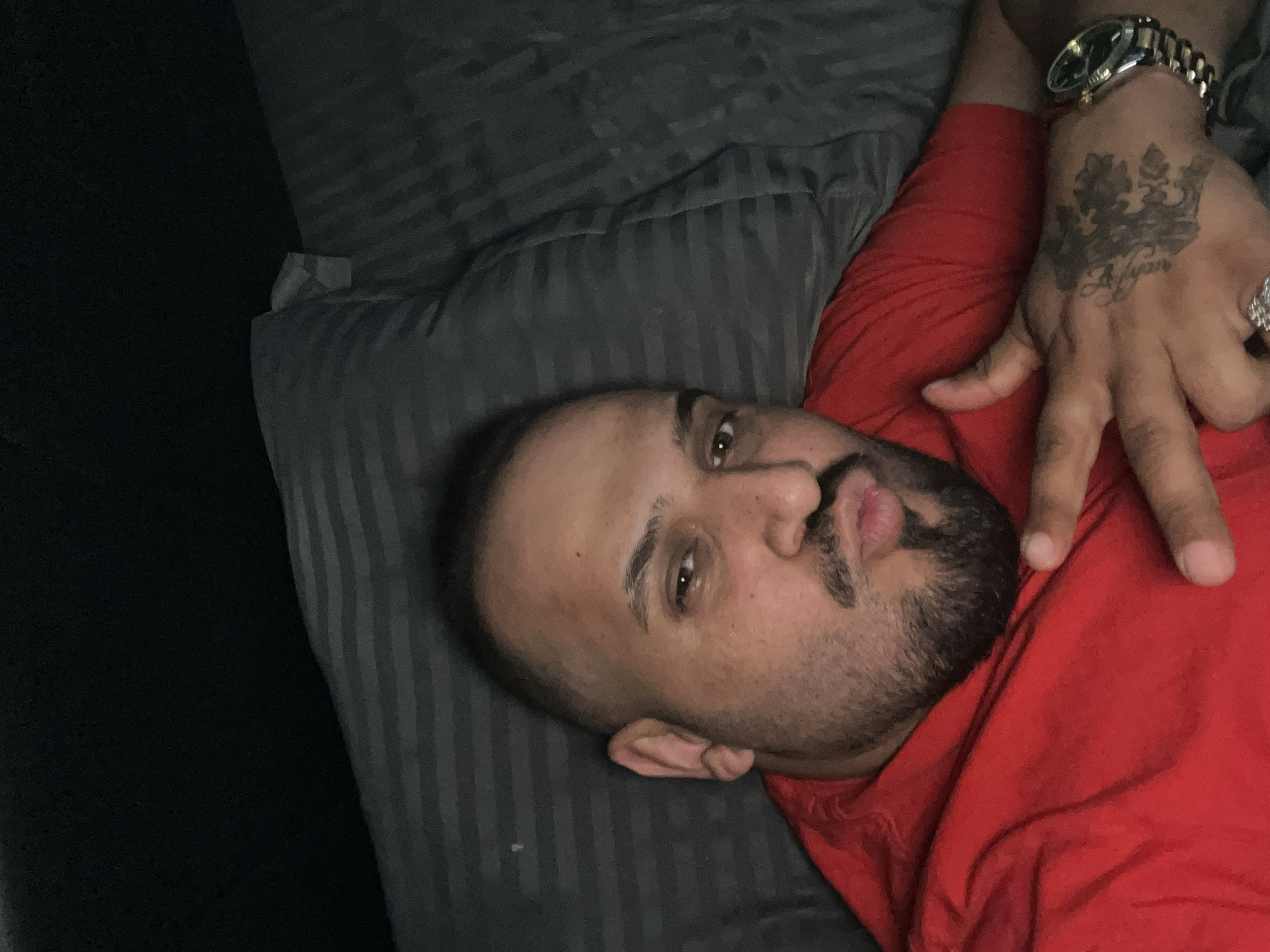 An adult male with light to medium skin tone, a shaved head, and a trimmed beard and mustache is depicted lying down, likely on a bed or couch. He is looking directly at the camera with a slightly puckered lip expression.
The man is wearing a red t-shirt. His left arm is raised, and his hand forms a "V" sign with his index and middle fingers, while his other fingers are bent. On his left wrist, he wears a prominent silver-colored watch with a metallic link strap. Several rings are visible on his fingers, including a large, possibly diamond-encrusted ring on his ring finger. A distinct tattoo on the back of his left hand features a crown design with the text "Alyan" written beneath it.
He is resting his head on a dark gray pillow with subtle vertical stripes. The area around him is dark and indistinct, suggesting an indoor setting with low lighting or a focus on the subject.