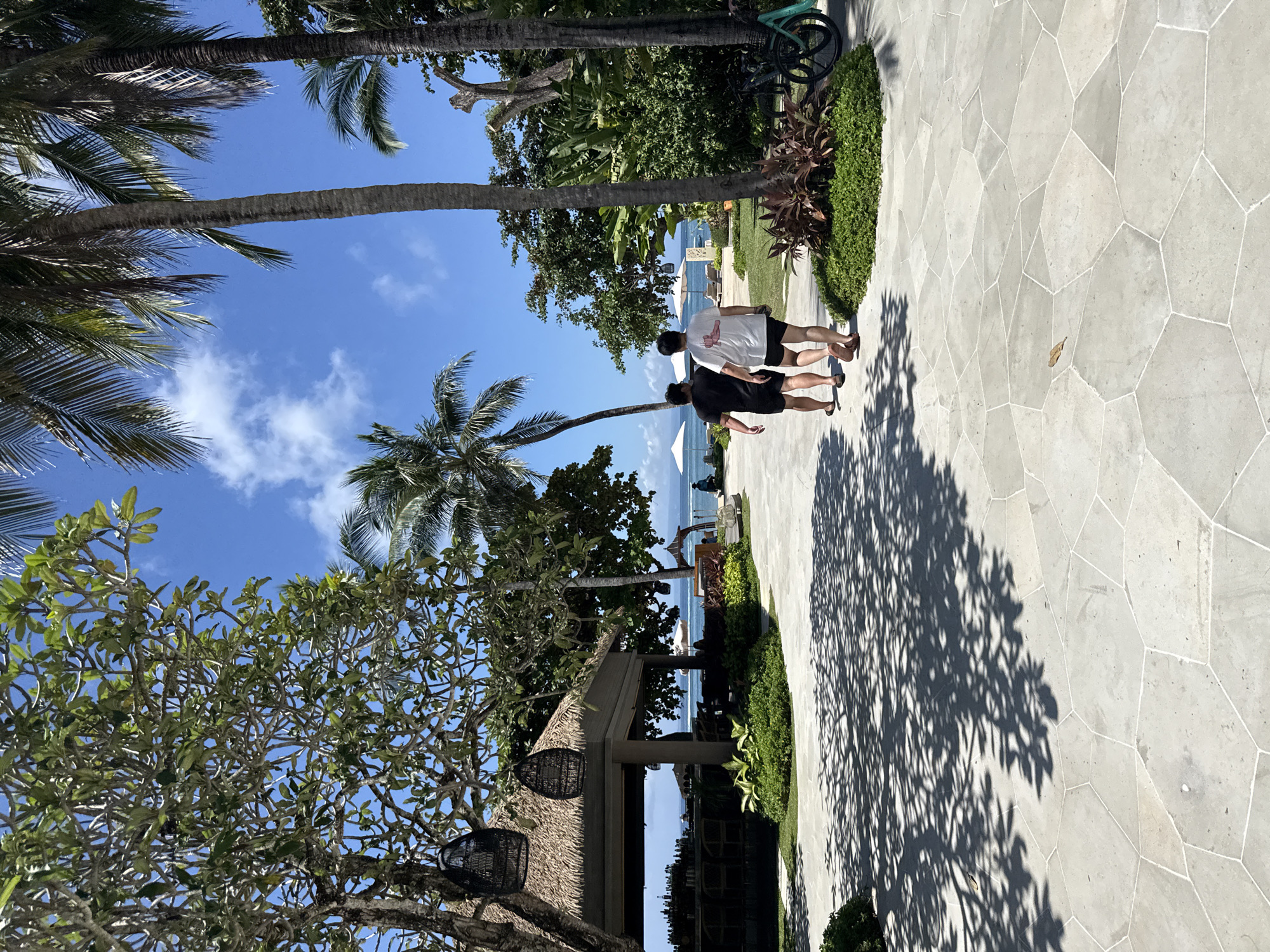 The image is a photograph taken in Nusa Dua, Indonesia. The scene depicts a paved walkway made of irregular stone slabs, flanked by lush tropical foliage including palm trees and other trees. Two individuals, dressed in casual clothing, are walking away from the camera along the path towards the ocean which is visible in the distance. Beach umbrellas are set up near the water. The angle of the sun is casting a large, detailed shadow of a tree onto the pathway. There are hanging basket-like decorative fixtures near a thatched roof structure. A bicycle is visible near the right upper corner of the scene. The sky is blue with scattered clouds.