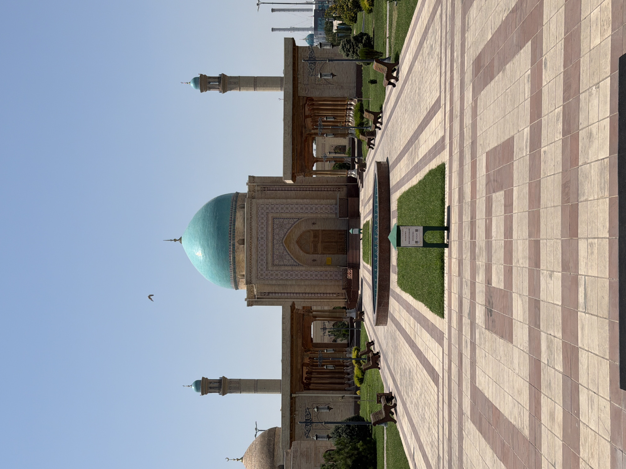 This image shows the exterior of the Hazrat Imam Mosque complex in Tashkent, Uzbekistan, under a clear blue sky. The main building features a large, vibrant turquoise dome and is flanked by two tall, slender minarets with similar turquoise caps. The mosque's facade is adorned with intricate geometric patterns and Arabic calligraphy, rendered in beige and brown tones.
In front of the mosque, a wide, paved walkway with a checkered pattern of beige and red-brown tiles leads towards the entrance. On either side of the walkway are manicured green lawns. Several wooden benches are strategically placed on the grass, facing the mosque. A small, rectangular fountain with turquoise water is visible in the mid-ground.
Further back, to the left of the main building, another dome, this one a lighter shade of beige, is partially visible. In the background, beyond the mosque complex, industrial structures including tall smokestacks can be seen, suggesting an urban or industrial area bordering the religious site. A single bird is captured in flight against the vast expanse of the sky. The scene is well-lit, indicating it was taken during daylight hours. There are no visible people in the image.
