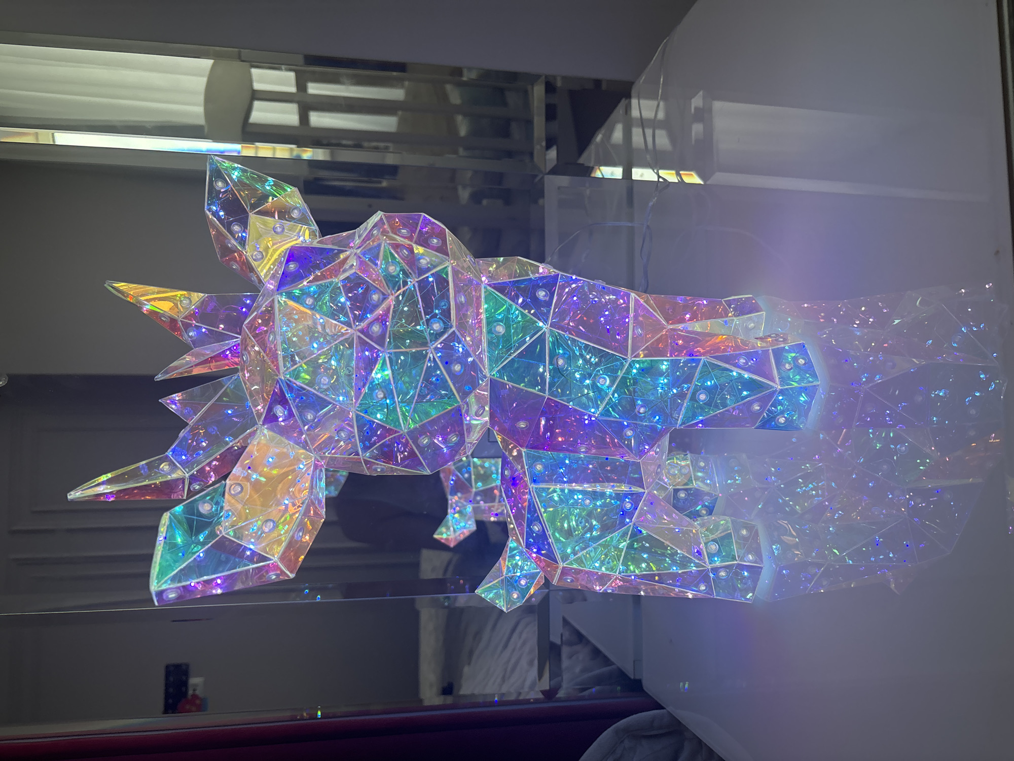 The image shows a geometric, iridescent light fixture with small embedded lights. The fixture is polygonal and reflects various colors. It is positioned in front of a mirrored surface, which creates multiple reflections of the fixture, including a fading effect on the right side. The background includes elements of a room, such as a wall, and possibly furniture. The location is Unknown, Unknown, as no specific details indicate the precise city or area.