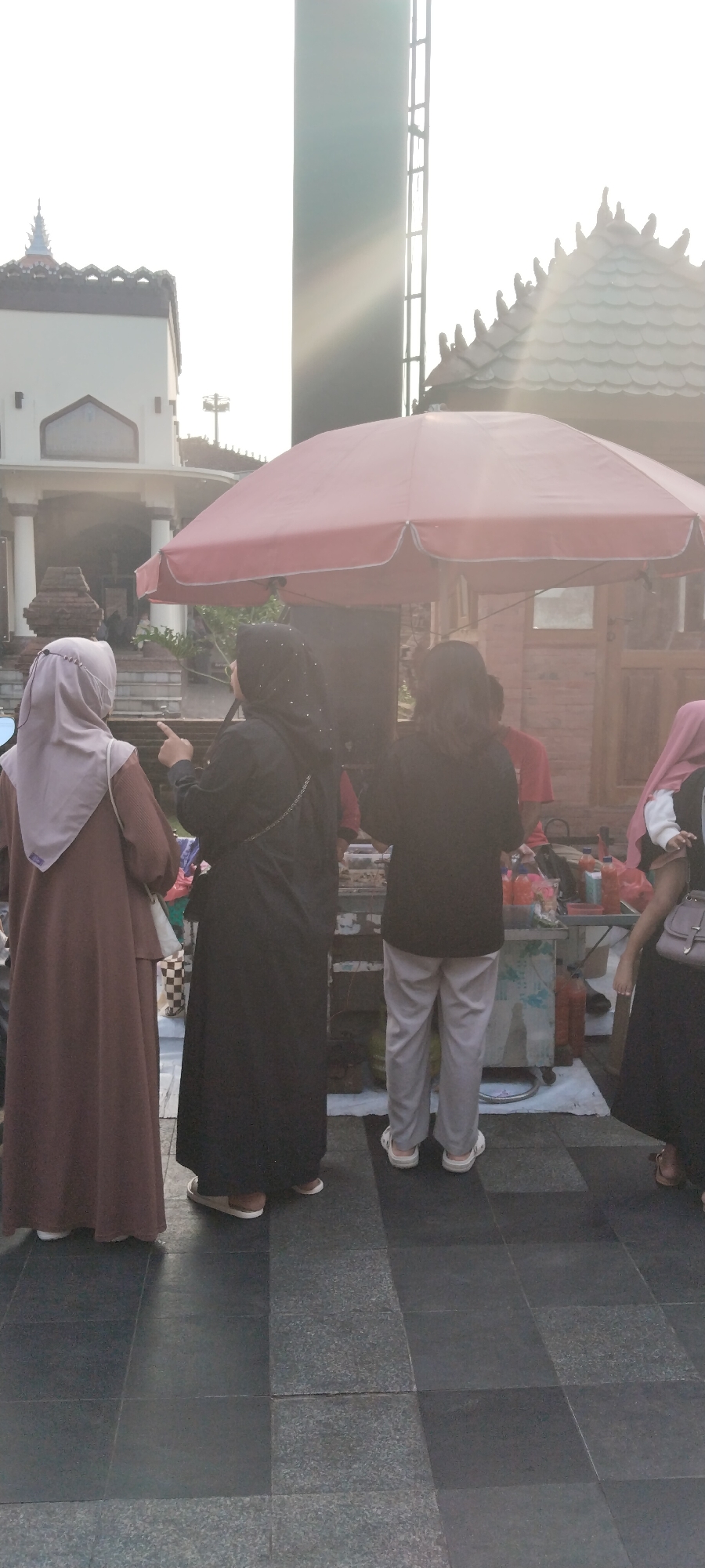 In the city of Kauman, Indonesia, a group of people stand near a street food stall under a large red umbrella. The sun is low in the sky, casting long shadows and creating a bright, hazy atmosphere. In the foreground, three women are visible from the waist down, two of whom are facing away from the camera, towards the stall. One woman, wearing a light brown hijab and a long brown dress, stands to the left. Next to her, a woman in a black hijab and black dress points towards something, possibly at the stall. To the right, a person in grey pants and a black top stands with their back to the camera. Behind them, a food cart with various bottles and containers is visible. In the background, traditional Indonesian architecture with distinctive tiled roofs can be seen, along with a tall, dark structure that resembles a tower or a part of a building. The ground is paved with dark grey tiles.