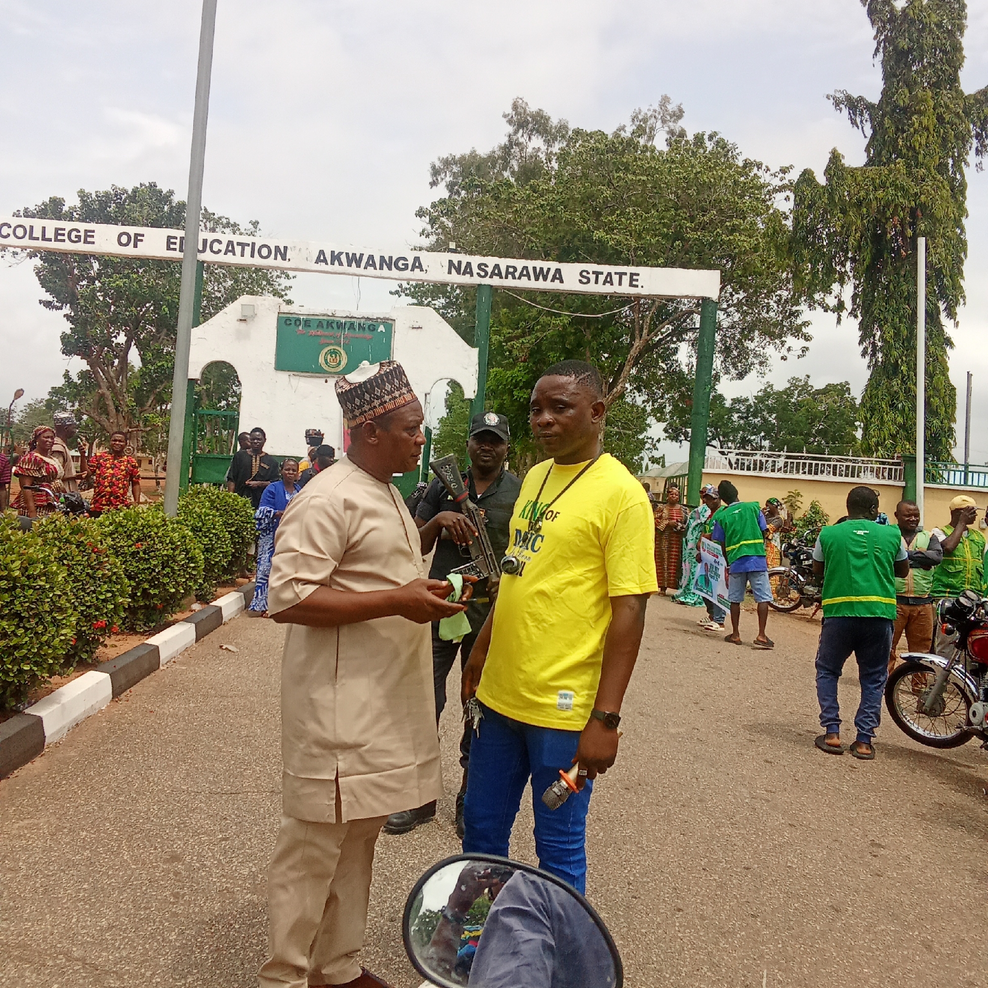 The image depicts an outdoor scene at the entrance of the College of Education, Akwanga, Nasarawa State, Nigeria. The large white gate in the background clearly displays the institution's name: "COLLEGE OF EDUCATION, AKWANGA, NASARAWA STATE." The sky is overcast, indicating it is daytime.
In the foreground, two men are conversing on a paved road. The man on the left is dressed in a light-colored traditional Nigerian attire with a patterned cap. He faces and appears to be speaking to the man on the right. This second man wears a bright yellow t-shirt emblazoned with the text "KING OF MIC" and blue jeans, holding a set of keys and a small metallic object, possibly a microphone head.
Behind them, a uniformed individual, likely a security guard, is visible holding a rifle. Scattered around the scene are several other people, suggesting a gathering or event. On the right side of the road, a group of individuals, some wearing green vests often associated with political or support groups, are standing near motorcycles. More people are visible on the left side, partially obscured by green bushes that line the road. The presence of a "KING OF MIC" t-shirt, a uniformed guard, and groups of people suggests an organized public gathering or event, possibly a political rally or community function.