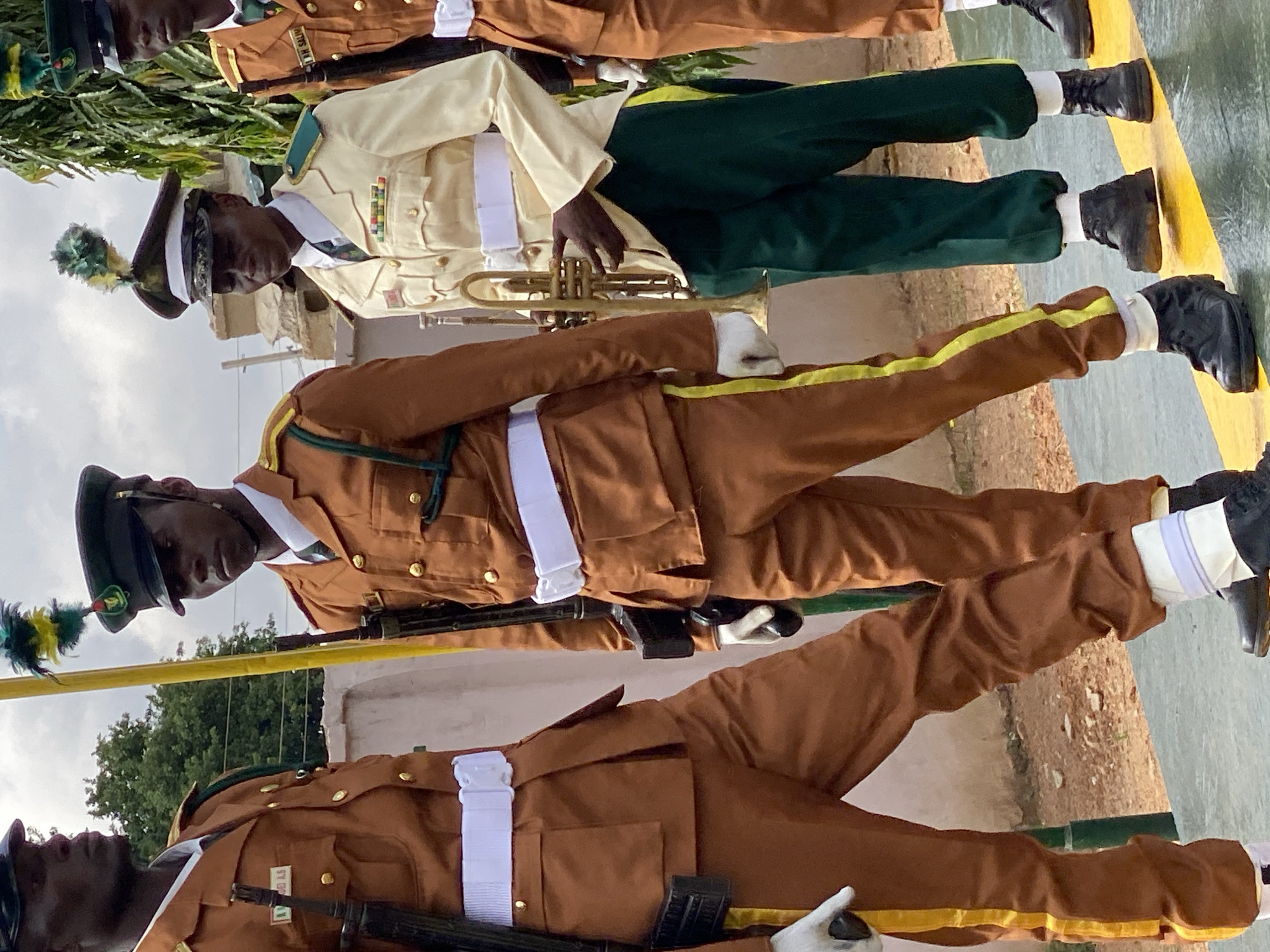 The image shows four individuals in military or ceremonial attire, standing in what appears to be an outdoor parade or formation. The individuals are wearing distinctive uniforms: two in a predominantly brown uniform with gold buttons and a yellow stripe down the side of their trousers, and two in a lighter colored uniform, one of whom is holding a trumpet. All individuals are wearing dark colored caps with feathered plumes. The background suggests an outdoor setting with some trees and buildings visible. The ground they are standing on is paved with a distinct yellow line running across it. Based on contextual information, this scene is located in Kaduna, Nigeria.