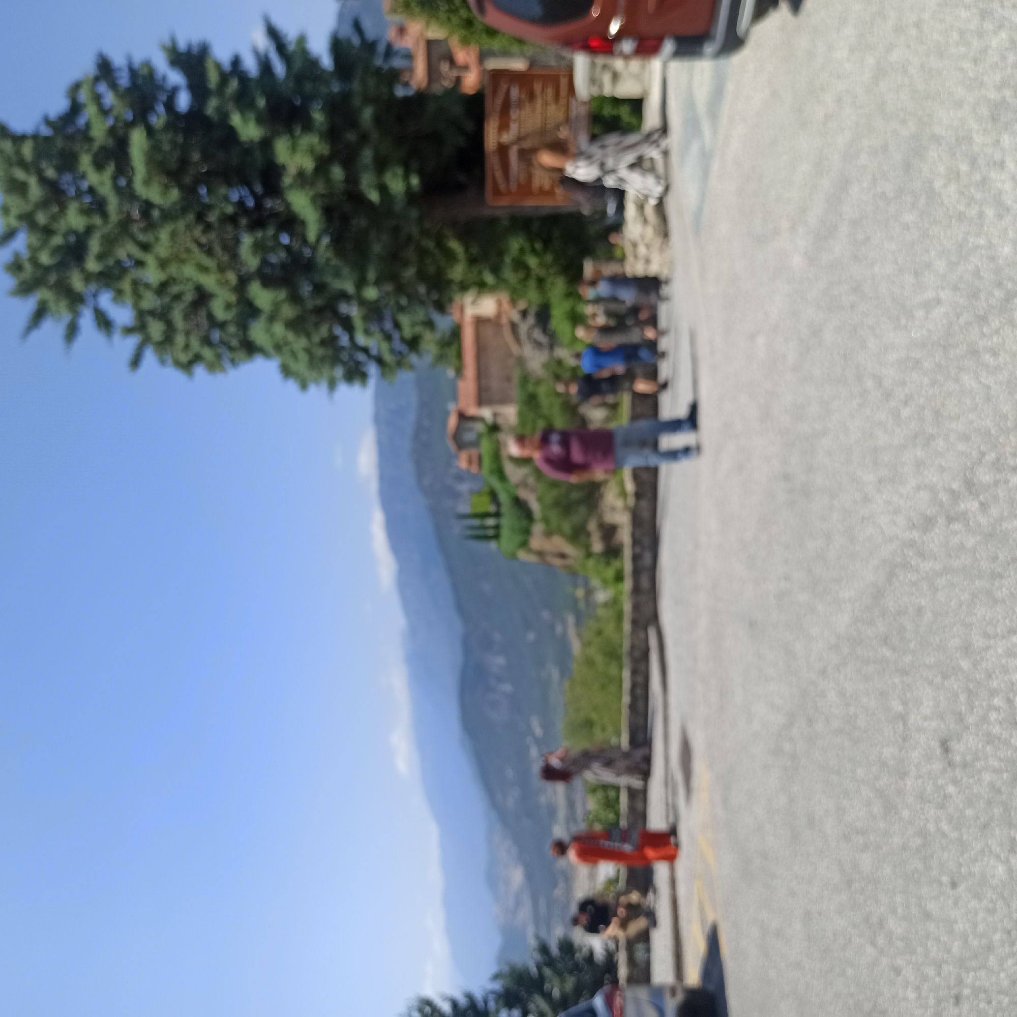 The image depicts a scene with several people in the foreground and a mountainous landscape in the background. The location is likely Meteora, near Kalambaka, Greece, as suggested by the distinctive rock formations and monasteries visible in the distance. In the immediate foreground, there is a paved area, possibly a parking lot or viewing area.
Several individuals are visible. To the left, a person wearing an orange dress stands facing away from the camera. Beside them, another person is seen from the back, wearing a patterned dress. Further to the left, a person in a black shirt and brown pants is seated. In the center of the image, a man in a maroon shirt and blue jeans stands facing left. To his right, a group of people are walking away from the camera, one wearing a blue shirt, and others in darker clothing.
In the background, the dramatic rock formations characteristic of Meteora rise up. Monasteries, with their distinctive reddish-brown roofs, are perched atop some of these rocks. The landscape is lush with green vegetation, and the sky is a clear blue. A large evergreen tree is prominent on the right side of the image, obscuring some of the background. A portion of a reddish-brown vehicle is visible on the far right edge of the frame. A wooden sign with text is also present in the mid-ground. The overall impression is of a popular tourist destination with historical and natural significance. The image is somewhat out of focus, suggesting it was taken with a camera that had difficulty achieving sharp focus in these conditions or at this distance.