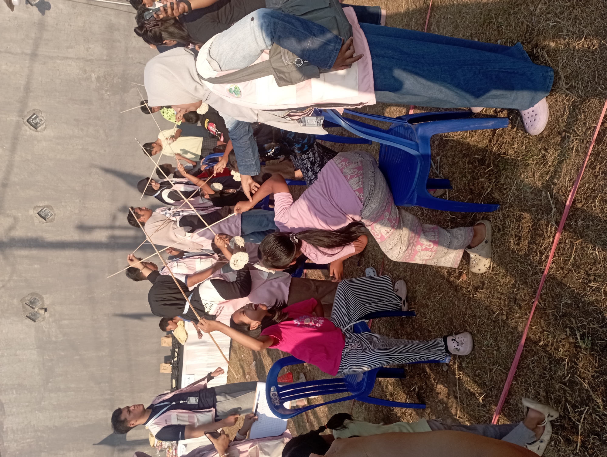 In Candirejo, Indonesia, on a sunny day, a group of people, predominantly children and young adults, are engaged in an outdoor activity in front of a grey concrete wall. The ground is covered with dry grass, and a pink string is laid out on it, suggesting a boundary or part of the game.
The central focus is a game involving long sticks, possibly chopsticks, used by participants to pick up small, white, circular objects. Two children in the foreground are particularly animated. One child, wearing a bright pink t-shirt and black and white striped pants, is seated on a blue plastic chair, laughing exuberantly while holding a stick with a white object. Another child, in a pink t-shirt and patterned pants, is also seated and actively participating.
Several other individuals are visible, some holding similar sticks and engaging in the game. A woman wearing a light blue hijab and a pink vest is standing and observing, holding a dark grey bag over her shoulder. Others are dressed in various casual attire, some wearing vests that appear to be part of an organizational uniform. In the background, a white tent and tables are set up, possibly for refreshments or as part of the event's setup. The lighting suggests it is either morning or late afternoon, casting shadows on the wall. The overall atmosphere appears to be one of cheerful participation and community engagement.