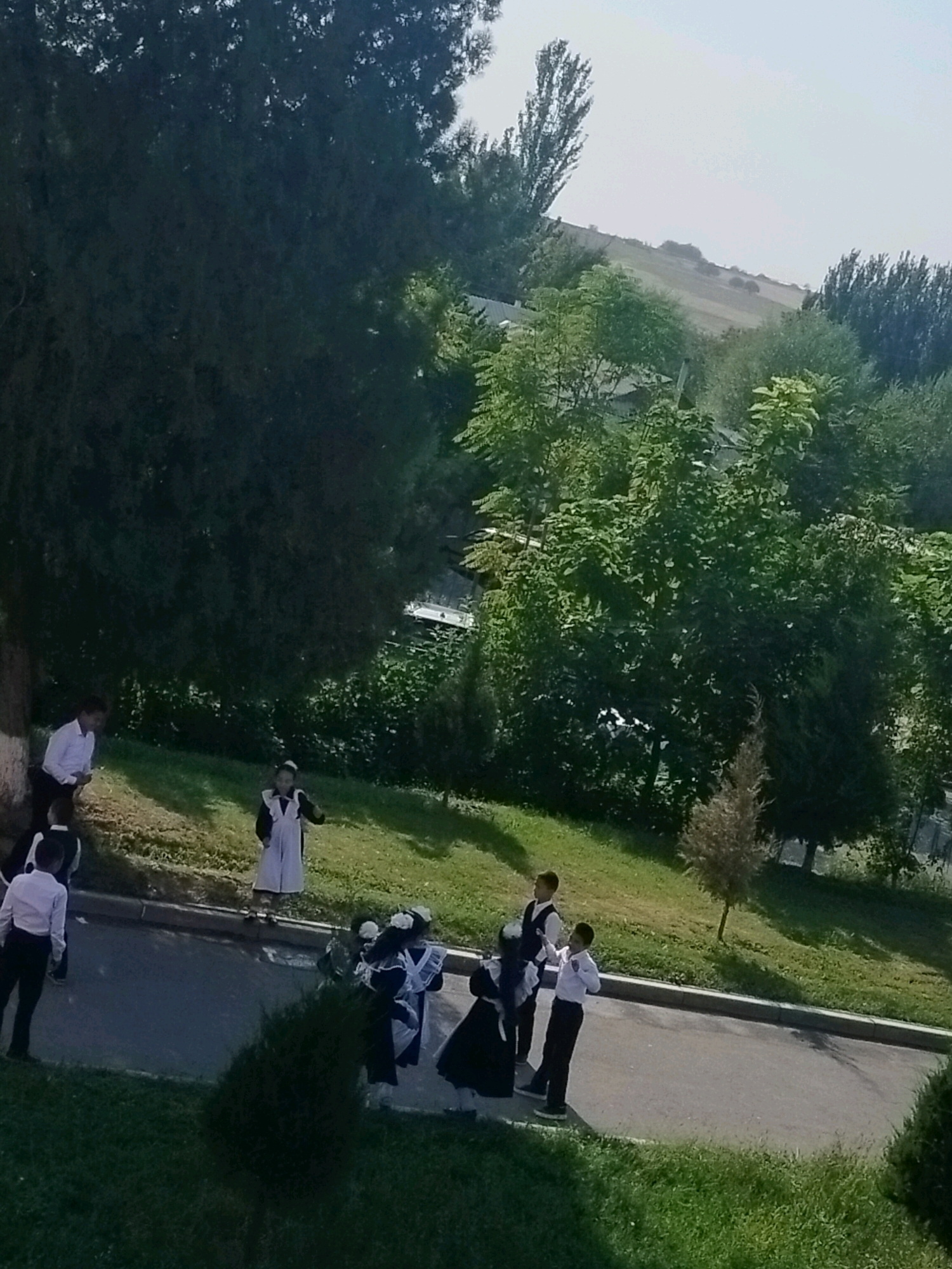 In an outdoor scene with a bright sky, several young people in school uniforms are gathered on a paved area and a grassy slope. On the paved area, three individuals are visible. One, wearing a white shirt and dark pants, stands facing away from the camera. Another, also in a white shirt and dark pants, is seen from the back, walking. To the right, a person in a white shirt and dark pants is standing and looking towards the right.
On the grassy slope above the paved area, several more individuals are present. One person, wearing a white dress and dark apron, is in the center, appearing to gesture. To their left, a person in a white shirt and dark pants stands. Further to the left and partially obscured, another person in a similar uniform is visible. In the foreground, a group of four individuals in black dresses with white aprons are clustered together. One of these is looking to the right, while another is facing away.
The environment includes lush green grass, mature trees with dense foliage, and a gently sloping, barren hillside in the background under a hazy sky. The scene suggests a school environment, possibly during a break or a departure. The presence of school uniforms and the layout of the grounds indicate an educational institution located in the city of Көлкент, Kazakhstan.