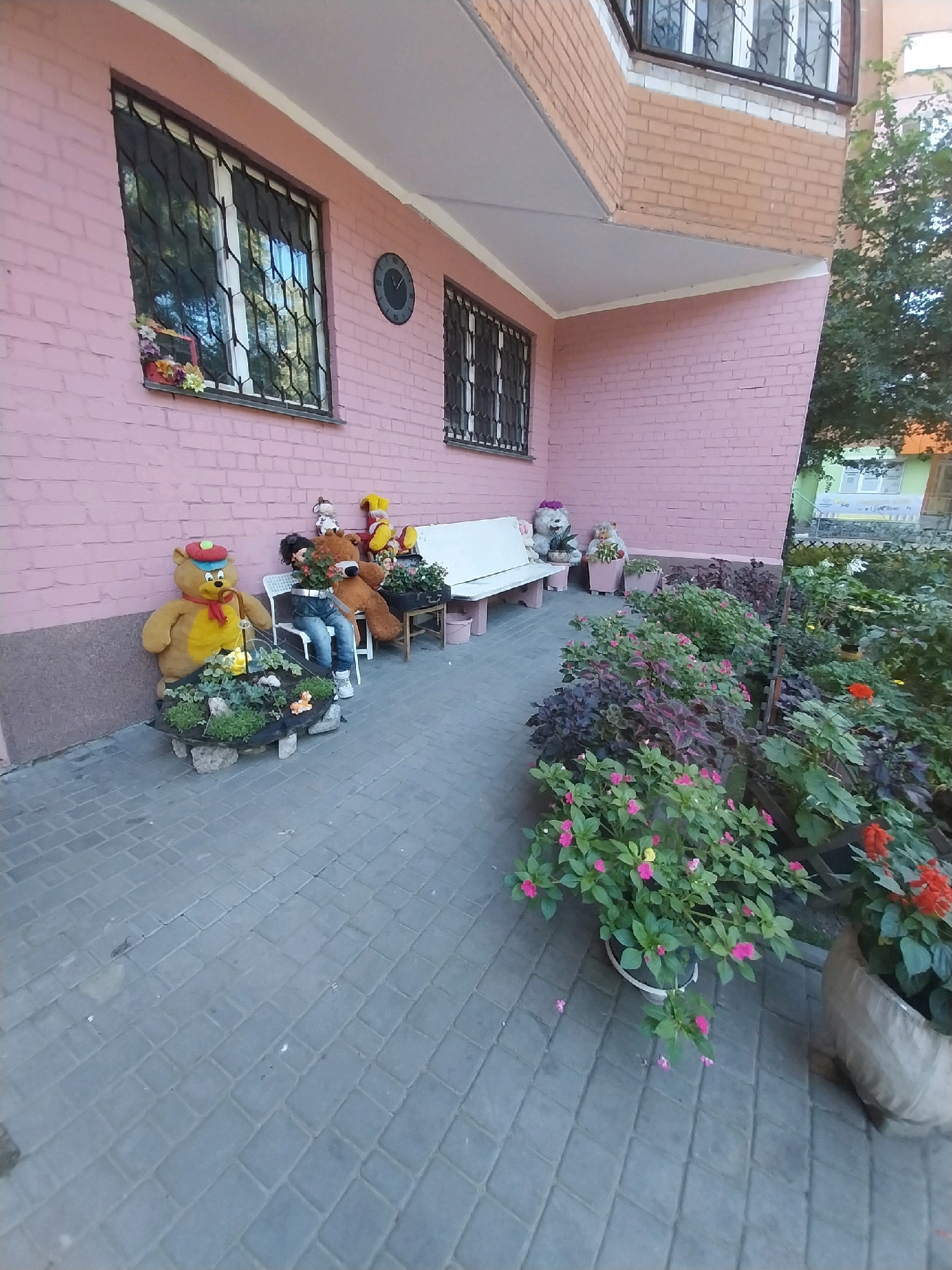 A low-angle, wide shot captures a section of a building's facade and an adjacent pathway in Dnipro, Ukraine. The building, painted a light pink, features barred windows. To the left, a window is adorned with a small arrangement of colorful flowers. A round clock is visible on the wall near another window.
In front of the building, a paved walkway is laid with grey interlocking pavers. Along the building's edge, a white bench is situated, flanked by two large stuffed teddy bears. One bear, wearing a red and blue cap and yellow scarf, sits in front of a planter box filled with small, green succulents and a few scattered pink flowers. Another, larger brown teddy bear is seated behind the planter, appearing to hold a bouquet of orange flowers. A smaller stuffed toy with a yellow head is also visible.
To the right of the teddy bears, a narrow path is bordered by a lush garden. The garden is densely planted with various flowers and foliage, including dark purple and green leafy plants, and vibrant pink and red flowers. Several potted plants are visible, including one large cloth sack planter holding bright red flowers. The scene suggests a residential area or a public space decorated with garden elements and stuffed toys. The lighting indicates it is daytime, possibly late afternoon or early morning due to the soft light.