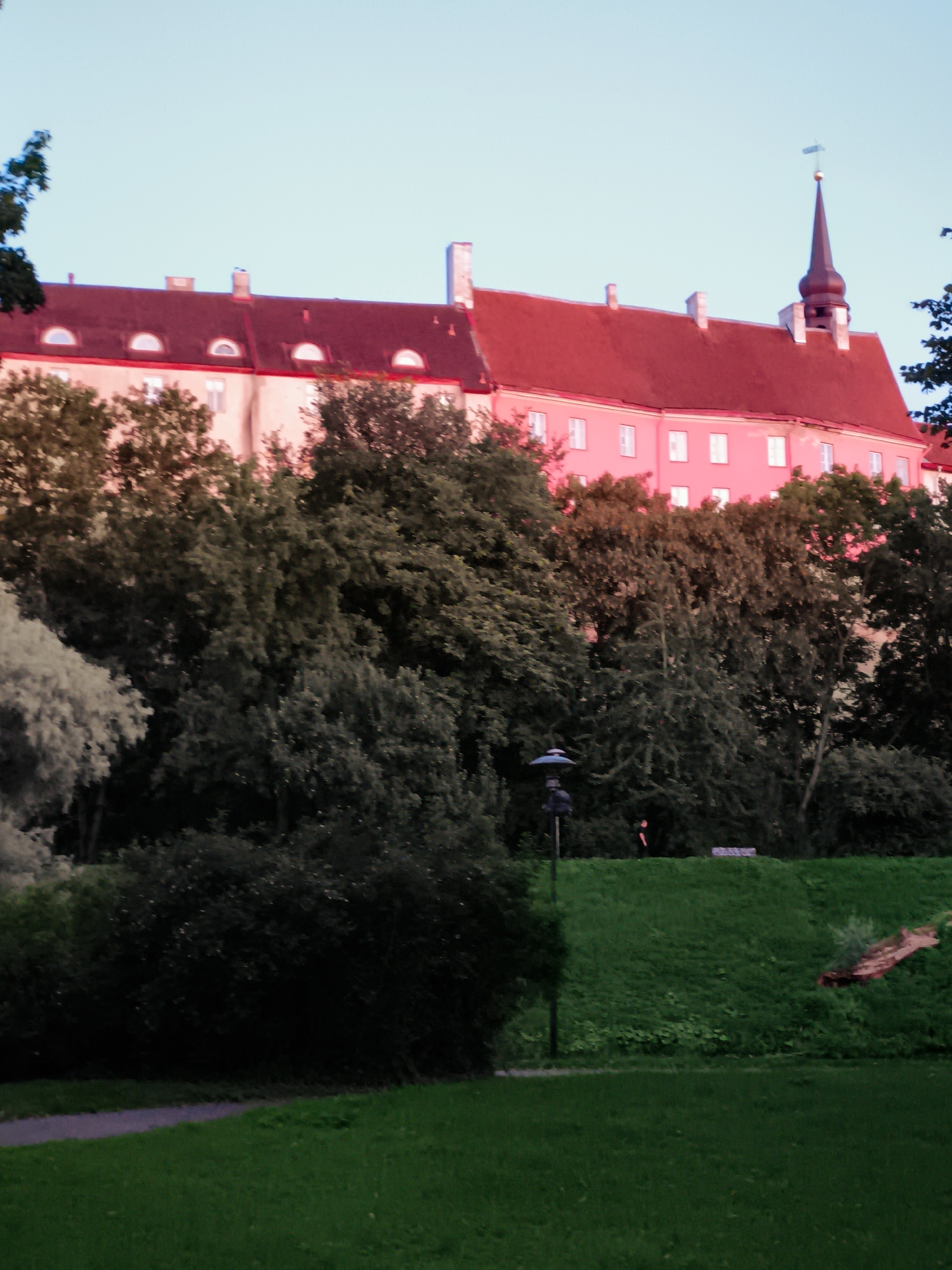 The image shows a building with a red roof in Tallinn, Estonia. The building appears to be historic, with a spire and several windows. The building is surrounded by trees, and there is a grassy hill in the foreground. A pathway is visible on the left side of the image.