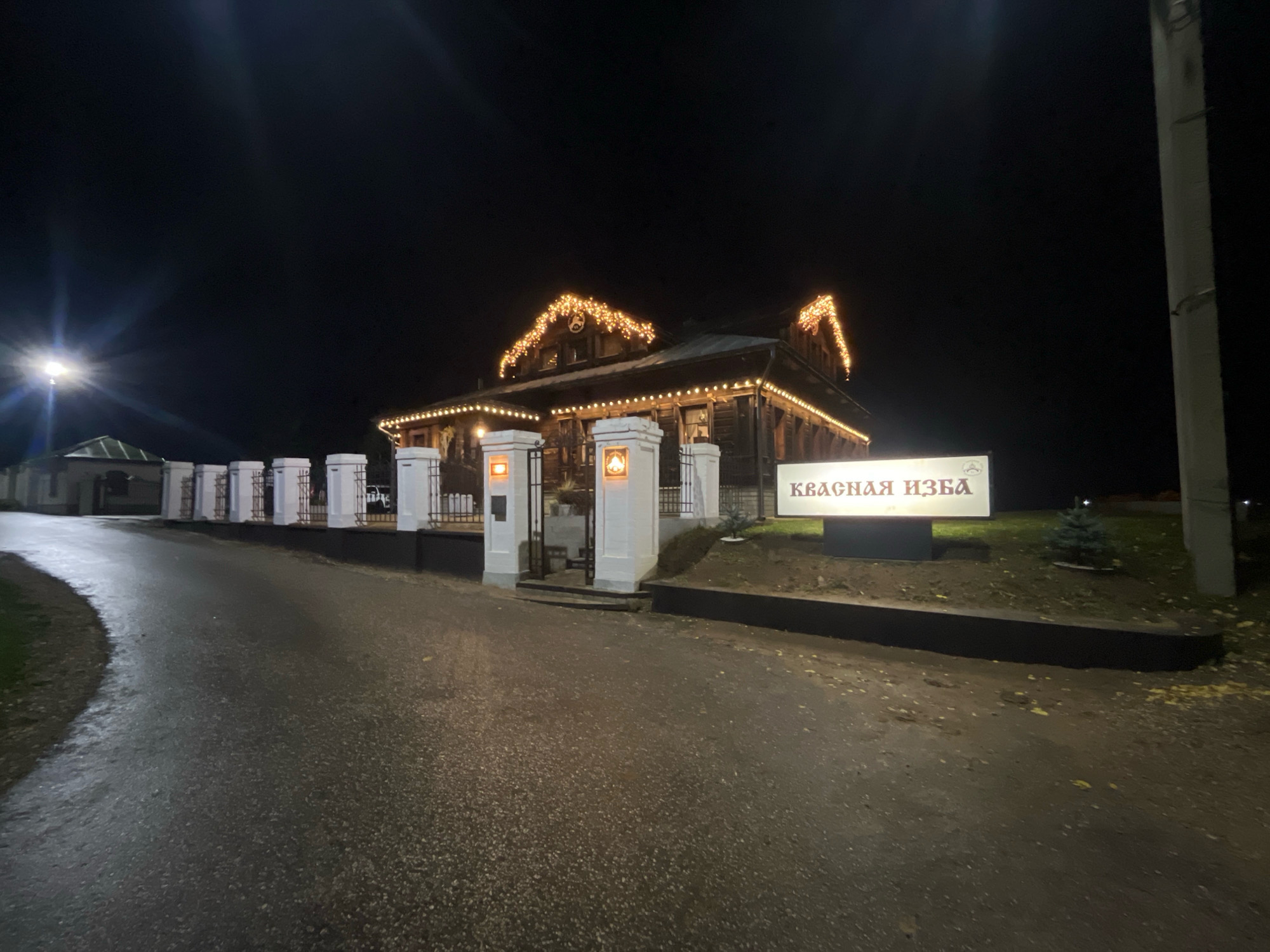 This is a night-time, eye-level, wide shot of "Krasnaya Izba," a log cabin restaurant located in Suzdal, Russia. The building is illuminated with festive golden string lights along its roofline and eaves. To the left of the building, a paved driveway curves away from the viewer. A row of white, ornate pillars with metal fencing separates the driveway from a grassy area in front of the building. A lit sign bearing the name "Krasnaya Izba" in Cyrillic is positioned to the right of the entrance. A small evergreen tree is planted in front of the sign. The ground is wet, suggesting recent rain. The sky is dark and clear.