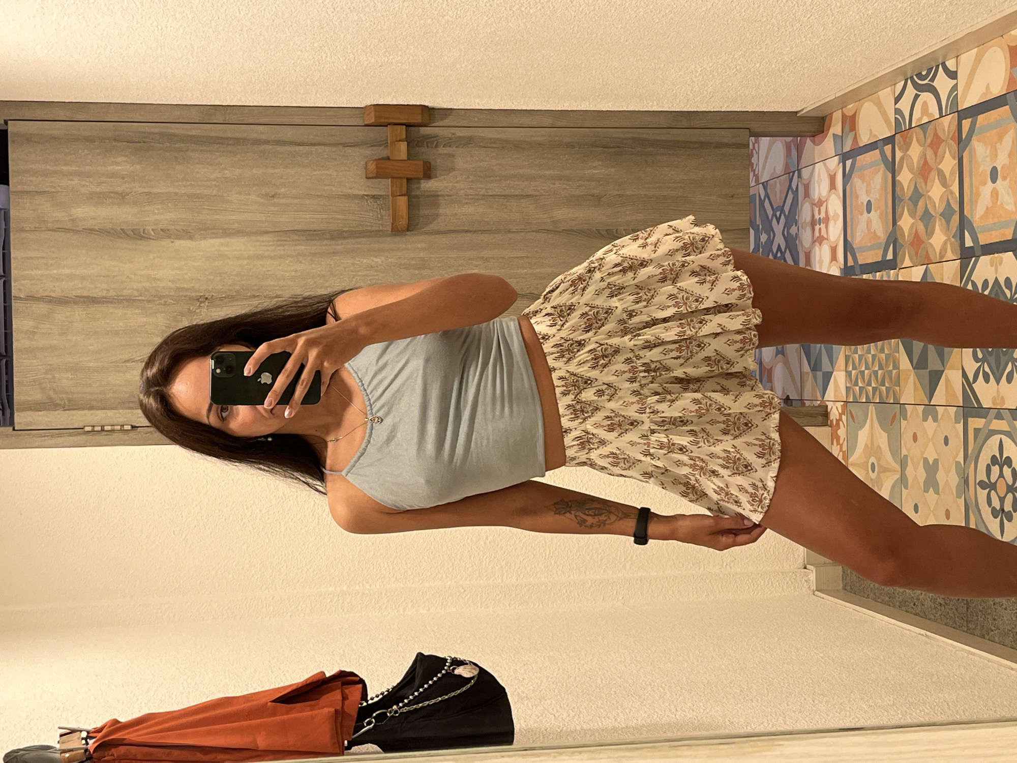 A young woman is taking a mirror selfie indoors. She has long, dark hair and tanned skin, wearing a light blue spaghetti strap crop top and a white mini skirt with a brown patterned design. A black fitness tracker or watch is on her left wrist, and a tattoo is visible on her left forearm. She holds a dark-colored smartphone, identified by the Apple logo on its back, covering part of her face as she captures her reflection.
The background of the indoor setting features light-colored wooden paneling behind her, adorned with a decorative wooden cross. To the right, a wall is covered in colorful, intricate patterned tiles in shades of blue, orange, and cream, suggesting a decorative or traditional aesthetic. The floor in the foreground is light-colored. In the bottom left reflection, parts of an orange and black bag are visible. The lighting indicates an indoor setting, potentially daytime or well-lit evening. The style of the patterned tiles could be a cultural cue for the Kuta Selatan, Indonesia location.