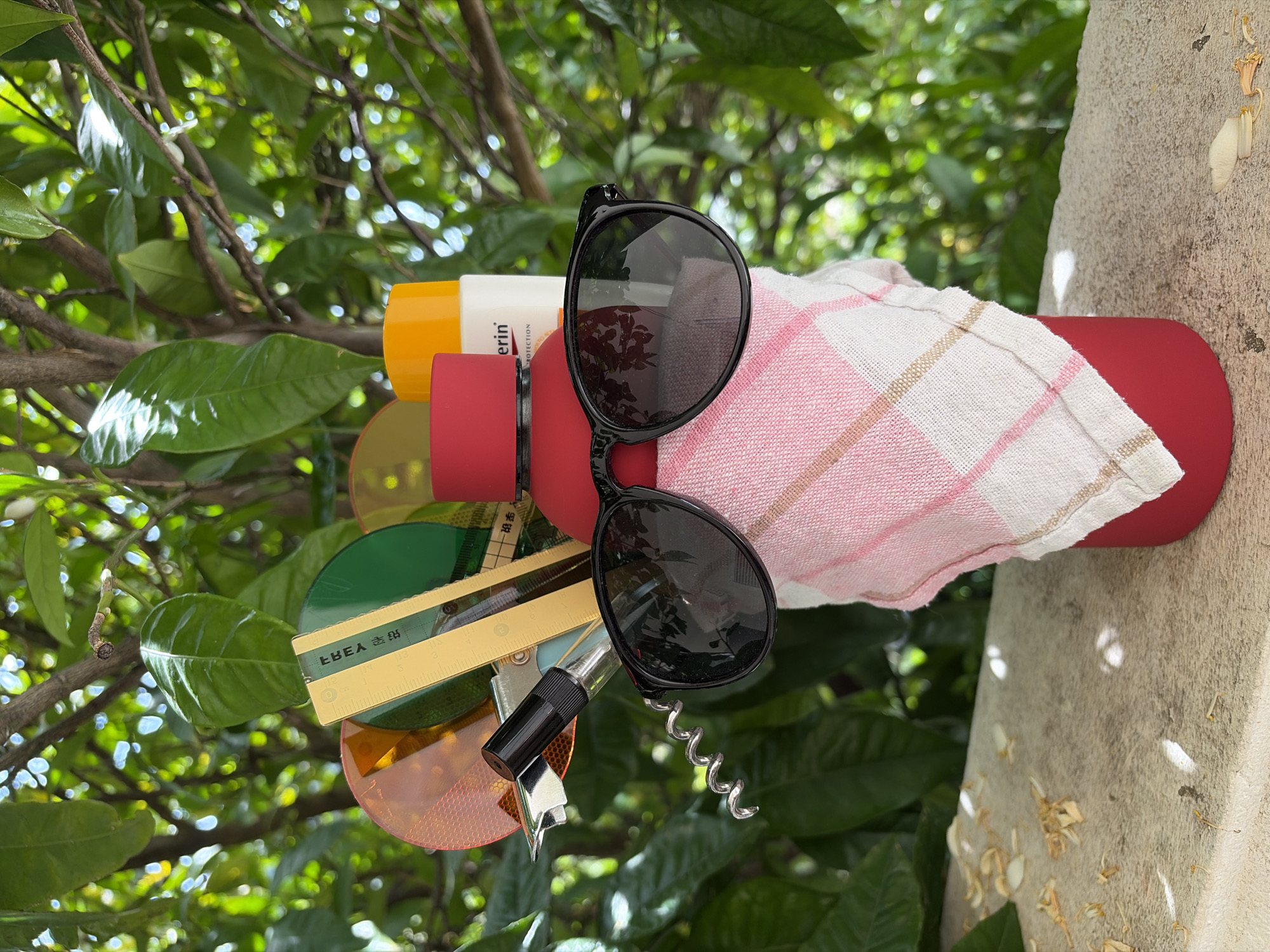 The media file displays a collection of personal items arranged outdoors, likely in a garden or on a balcony, with a lush green foliage background. The primary subject is a red cylindrical object, possibly a water bottle or thermos, wrapped in a pink and white checkered towel. Perched on top of the red bottle are a pair of black sunglasses with dark lenses.
To the left of the sunglasses, a white and yellow sunscreen bottle is visible, bearing the brand name "Eucerin" and the word "PROTECTION". Below these items, a variety of objects are clustered: several colorful circular filters (green, yellow, orange), a metallic ruler with "IKEA" text and measurement markings, and a corkscrew with a black handle. Another small, dark, possibly bottle opener tool is also present among these items.
The background features vibrant green leaves and branches, indicating a natural, outdoor setting. The light suggests it is daytime, with some bright highlights on the leaves and reflections on the sunglasses. To the right of the central arrangement, a textured concrete pillar or wall is visible, with some fallen white and yellow dried petals or flower debris scattered on its surface.
While no direct activity is taking place, the collection of items—sunscreen, sunglasses, water bottle, towel, and corkscrew—strongly suggests preparation for a leisurely outdoor activity, such as a picnic, beach trip, or a relaxed day in the sun, consistent with a holiday setting in Budva, Montenegro. The scene evokes a feeling of summer relaxation and enjoyment.