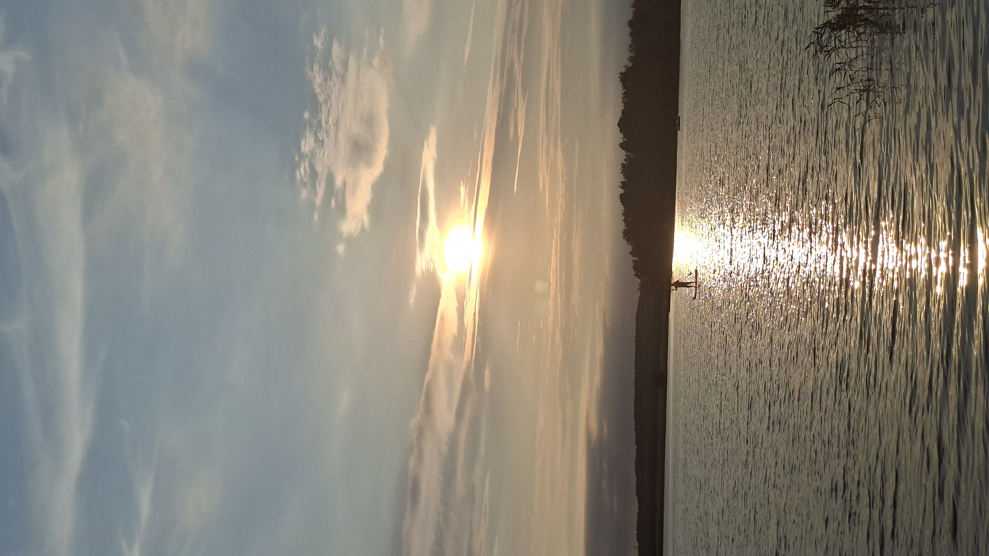 The image depicts a serene sunset over a body of water, likely a lake or river, with a single person paddleboarding in the distance. The sky is a gradient of soft blues and oranges, with wispy clouds catching the golden light of the setting sun. The sun itself is a bright, luminous orb positioned in the upper half of the frame, casting a shimmering reflection across the water's surface. In the foreground, the water is calm with gentle ripples, and reeds are visible along the right edge of the frame. A dark, silhouetted treeline forms the horizon in the background. The overall atmosphere is tranquil and peaceful. The location is identified as Smalyavichy, Belarus.