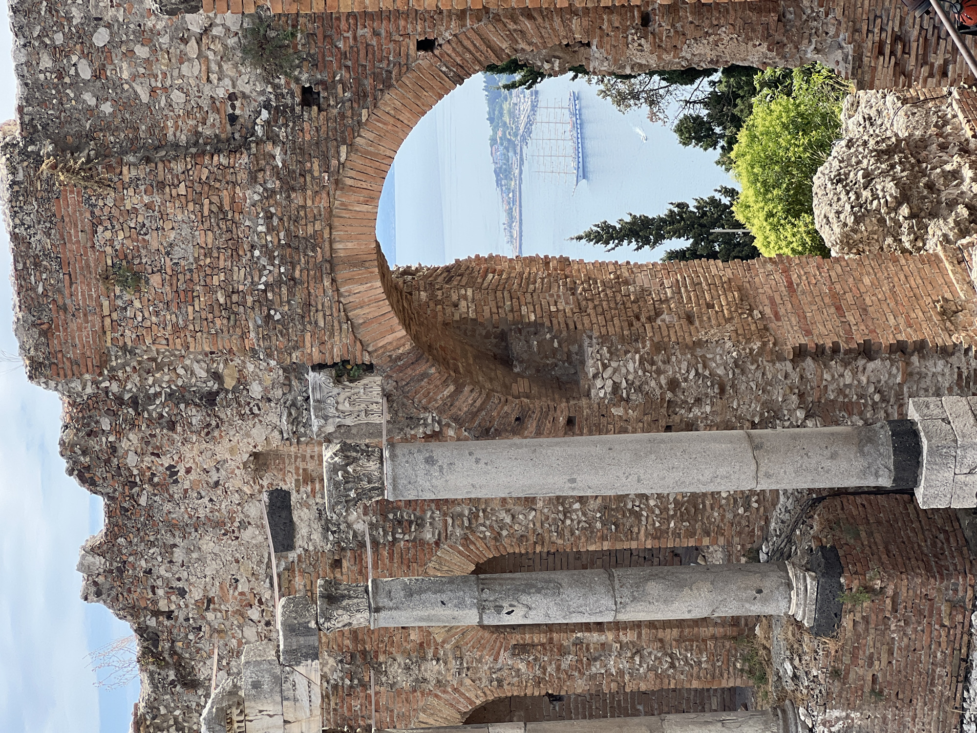 The image shows a view through an arched opening in the ruins of an ancient theater in Taormina, Italy. The theater is constructed of brick and stone, with several large stone columns visible in the foreground. Through the arch, a scenic vista of the sea unfolds, featuring a large sailing ship with multiple masts. The water is calm, and the sky is a clear blue with some scattered clouds. Trees and vegetation are visible on the coastline in the distance, along with buildings that suggest a town or city. The overall impression is one of historical grandeur and natural beauty.