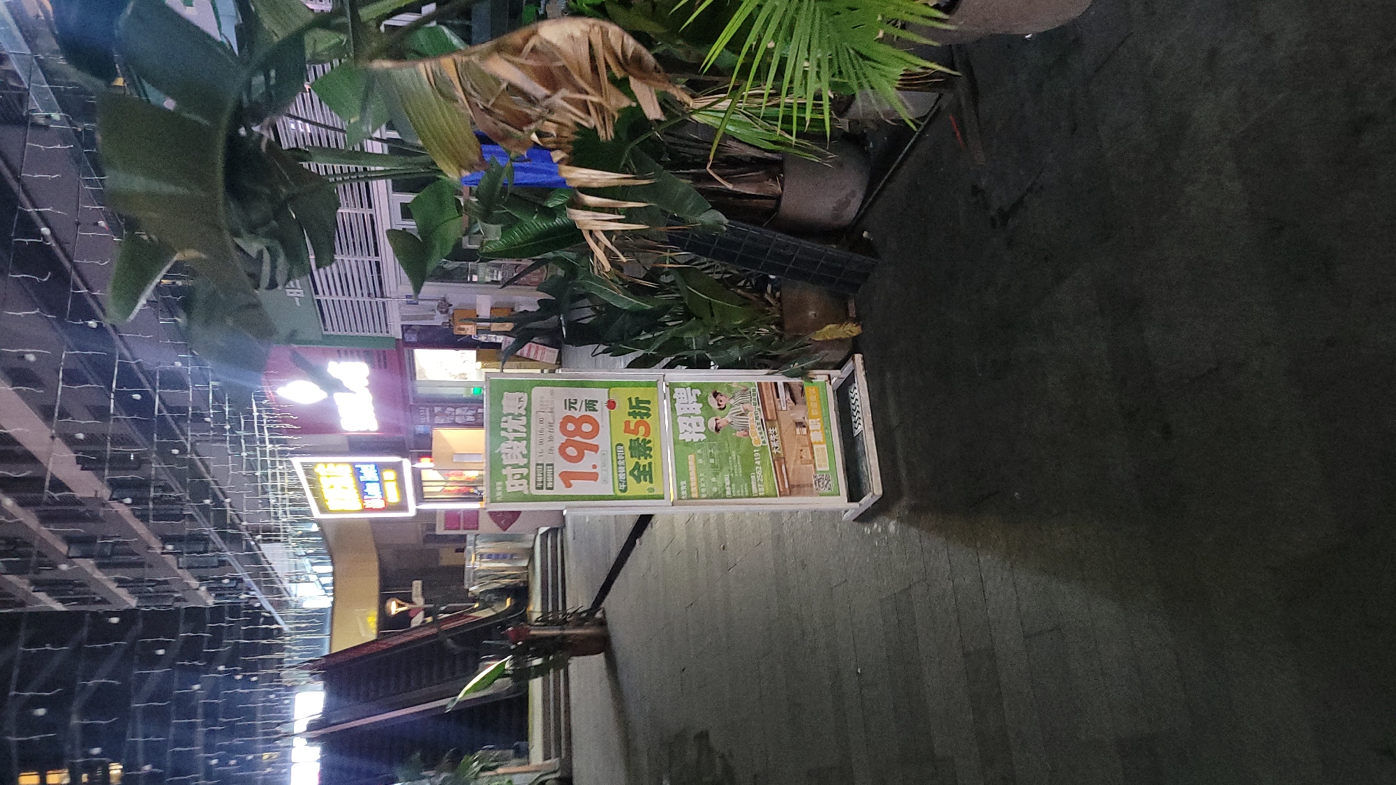 The image depicts a street scene in Jiulongpo District, Chongqing, China. A sign advertises a special deal of 1.98 yuan for a limited time, with an additional 50% off vegetarian options. Another sign is advertising job openings. The environment appears to be urban, as suggested by the street lighting and infrastructure, with plants and greenery around.