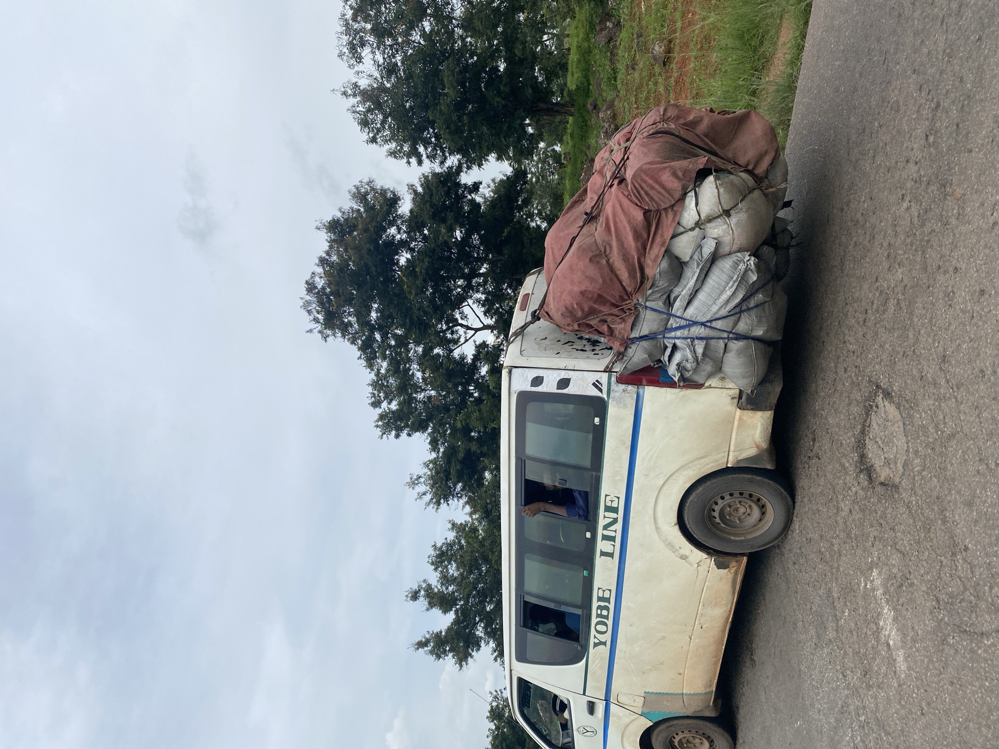 A white van with "YOBE LINE" printed in blue lettering along its side is parked on a cracked asphalt road, with several large bags of goods secured to its rear. The van is dirty and shows signs of wear, including rust and scuff marks. The bags are covered with a reddish-brown tarp and tied with rope. In the background, several large trees with dark green foliage are visible against a cloudy sky. The scene appears to be in a rural or semi-rural area outside of Makera, Nigeria. One passenger is visible through the van's window, with their arm raised.