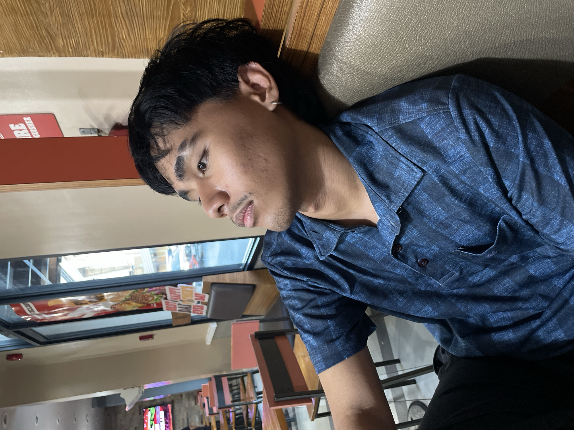 A young Filipino male with short, dark hair and a small mustache is pictured in profile, looking upwards and leaning against a wooden wall. He is wearing a dark blue, plaid short-sleeved shirt and dark pants. A silver hoop earring is visible in his left ear. The background suggests an indoor dining area, possibly a fast-food restaurant, with tables and chairs visible. In the distance, through a glass window, a street scene with vehicles can be seen. A red sign with the word "FIRE" is mounted on the wall behind the man. The lighting is dim, with a warm tone. The location is likely a commercial establishment within Davao City, Philippines, given the subject's ethnicity and the commonality of such establishments in urban areas of the Philippines.