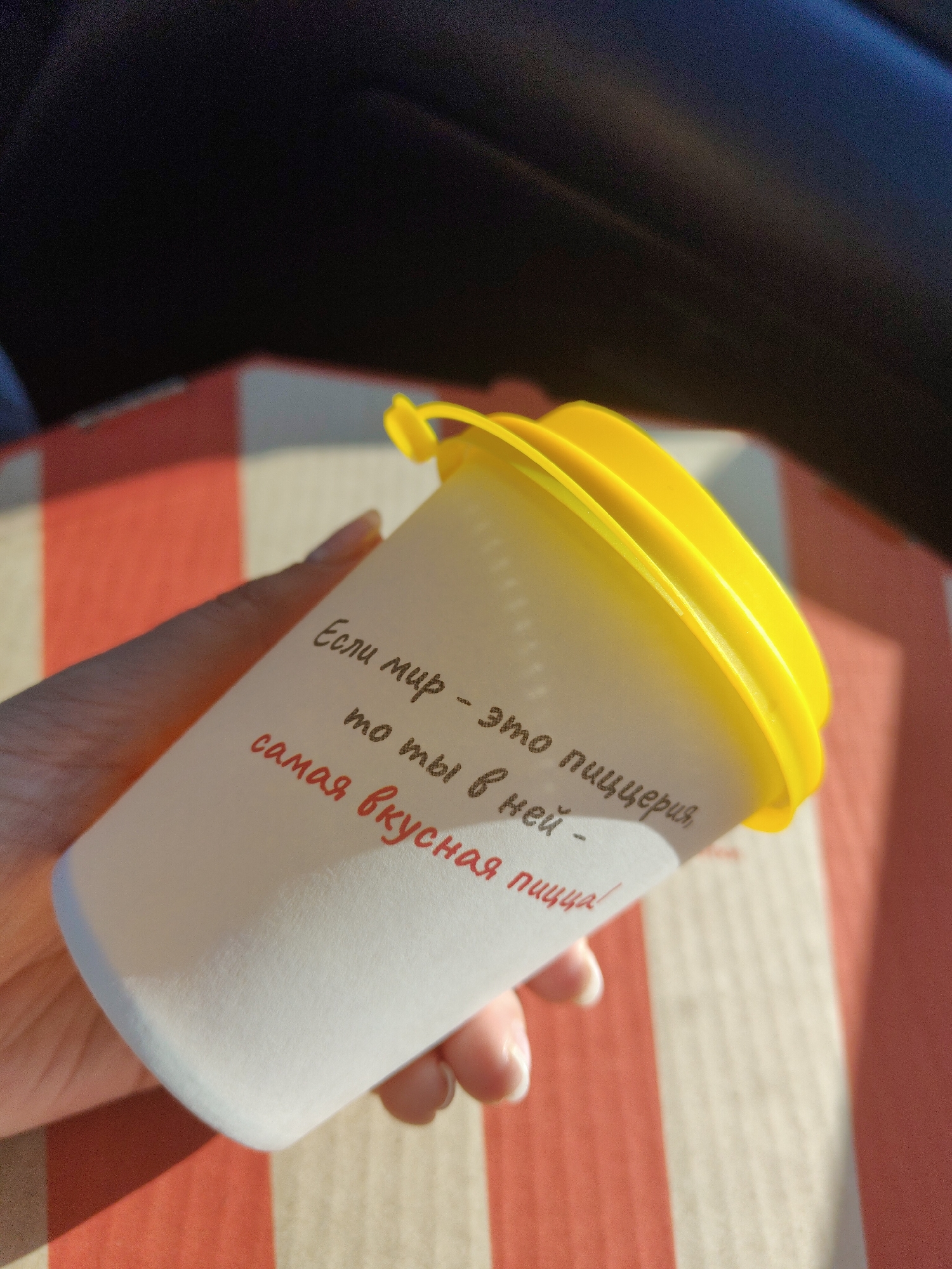 A fair-skinned hand holds a white paper cup with a yellow lid. The cup is angled towards the camera and rests on a surface with alternating red and white stripes. The text on the cup, written in a red-brown color, states "Если мир - это пиццерия, то ты в ней - самая вкусная пицца!", which translates to "If the world is a pizzeria, then you are the most delicious pizza in it!". The background is slightly blurred, but there's a dark area visible at the top, possibly indicating an interior setting.