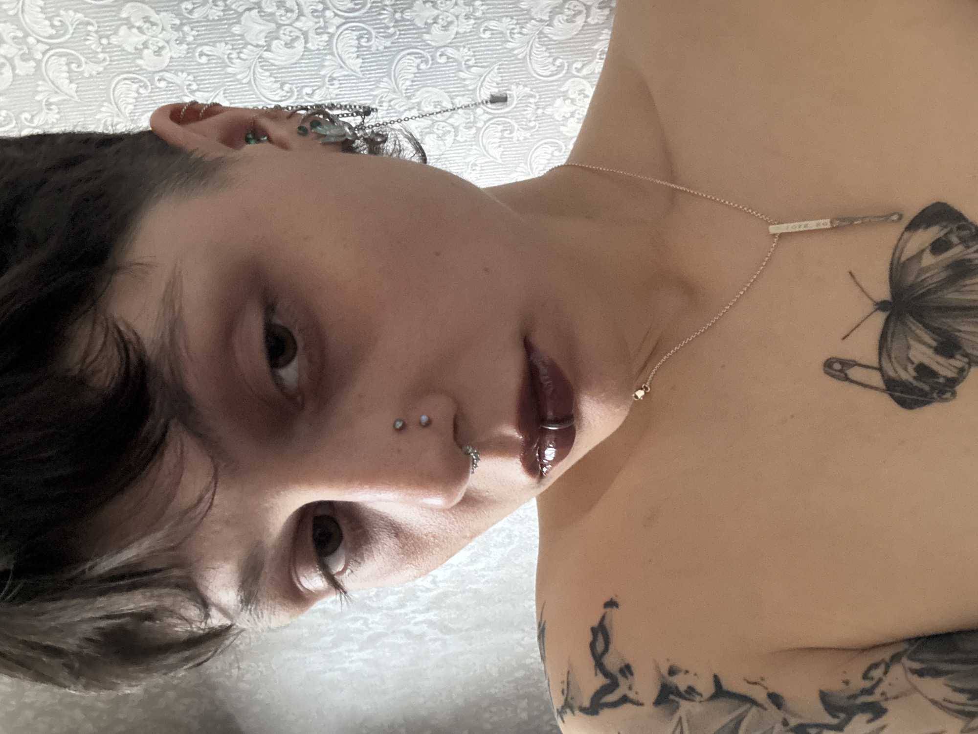 The image is a close-up portrait of a person with multiple piercings in their ears and nose. They have dark hair, fair skin, and brown eyes. A delicate necklace with a bar pendant rests on their chest, near a tattoo of a butterfly with a safety pin. Additional tattoos are visible on the shoulder and upper chest. The background features a patterned white surface. There is no indication of Saint Petersburg, Russia.