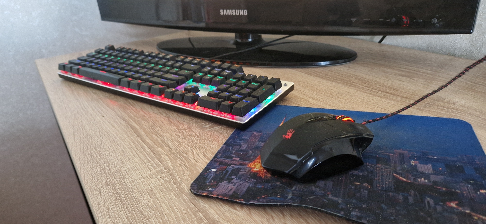 This media file displays a close-up, eye-level shot of a gaming setup on a wooden desk. The primary objects are a black mechanical keyboard with RGB backlighting and a black gaming mouse resting on a mousepad. The keyboard exhibits a variety of colors from its illuminated keys, including red, green, blue, and yellow. The mouse also has illuminated accents, appearing orange. The mousepad features a nighttime cityscape scene, possibly a famous landmark like the Eiffel Tower in Paris.
Behind the keyboard and mouse, a portion of a black Samsung computer monitor is visible. The monitor has a curved stand and displays some icons on its lower bezel, including a red illuminated logo that resembles a silhouette of a person. The overall scene is indoors, suggesting a personal computer setup. The lighting appears to be a mix of ambient room light and the artificial glow from the computer peripherals. There are no people visible in the frame. The desk surface has a light wood grain texture. The image is sharp and focused on the keyboard and mouse.