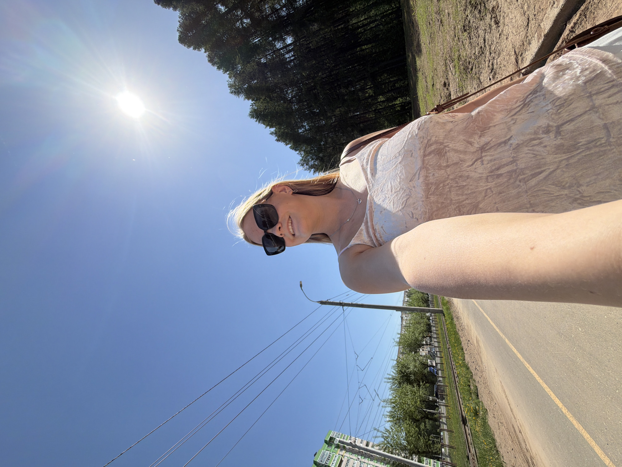 A woman with blonde hair and sunglasses is visible in the frame, wearing a light-colored dress. She is positioned near a street or path marked with a yellow line. Power lines and utility poles are visible against a bright blue sky with the sun shining brightly. In the background, there is a view of buildings and trees, with a dense forest area further in the distance. The location appears to be in the city of Izhevsk, Russia.
