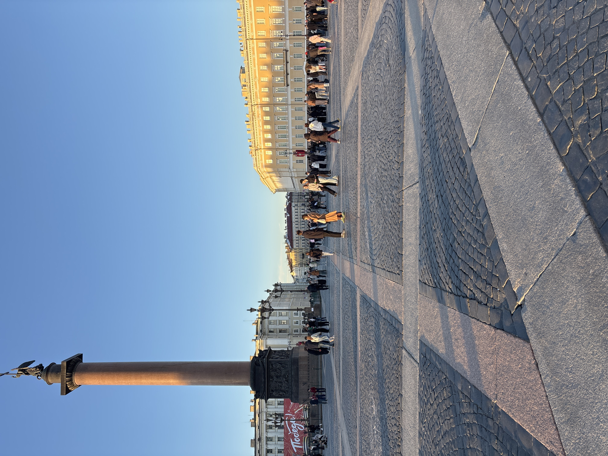 The image shows Palace Square in Saint Petersburg, Russia. A tall, smooth, brown-colored pillar, adorned with a dark sculpture at its peak, stands against a clear blue sky on the left side of the image. To the right, buildings line the square. People are present in the square, walking near and in front of the buildings. The ground is paved with a mixture of gray stone slabs and smaller, dark-colored cobblestones. The image is taken in daylight, with sunlight casting shadows on the ground and buildings.