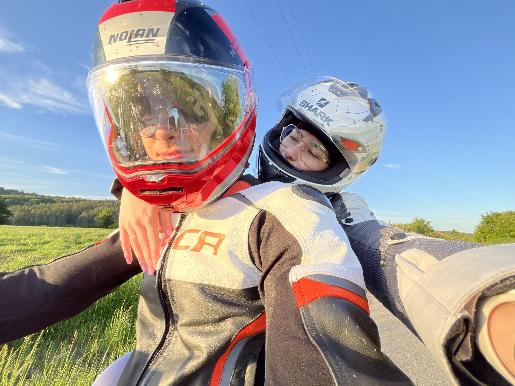 Two individuals, wearing full-face motorcycle helmets, are posing for a selfie outdoors. The person on the left wears a red, white, and black helmet with the brand "NOLAN" visible. Their helmet visor is heavily covered with insect debris. They wear glasses and red lipstick, and their left arm is extended, with a hand resting on the other person's shoulder. Their hand is manicured with light pink nail polish and displays a ring on the ring finger. They are wearing a black, white, and gray motorcycle jacket with red accents, featuring the letters "CA" on the white portion. The person on the right wears a white and gray helmet with the brand "SHARK" visible. They also wear glasses. Their torso is positioned behind the other person. They wear a gray and white motorcycle jacket. The background consists of a grassy field leading to trees in the distance and a clear blue sky with power lines visible. The location appears to be a roadside or scenic overlook.