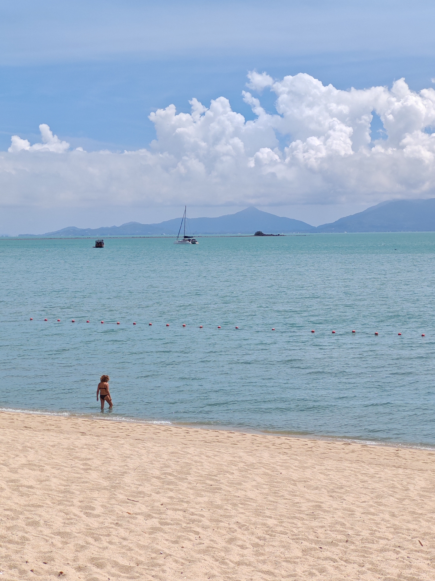 A person stands in shallow, turquoise water at the edge of a sandy beach, facing away from the camera. The person is wearing a dark-colored bikini. A string of red buoys floats on the water's surface, marking a boundary. In the distance, a sailboat with a white sail is visible, along with a smaller boat. Beyond the water, a range of hazy, blue mountains forms the horizon. The sky above is a bright blue, dotted with fluffy white clouds. The scene appears to be a coastal area in Baan Bo Phut, Thailand.