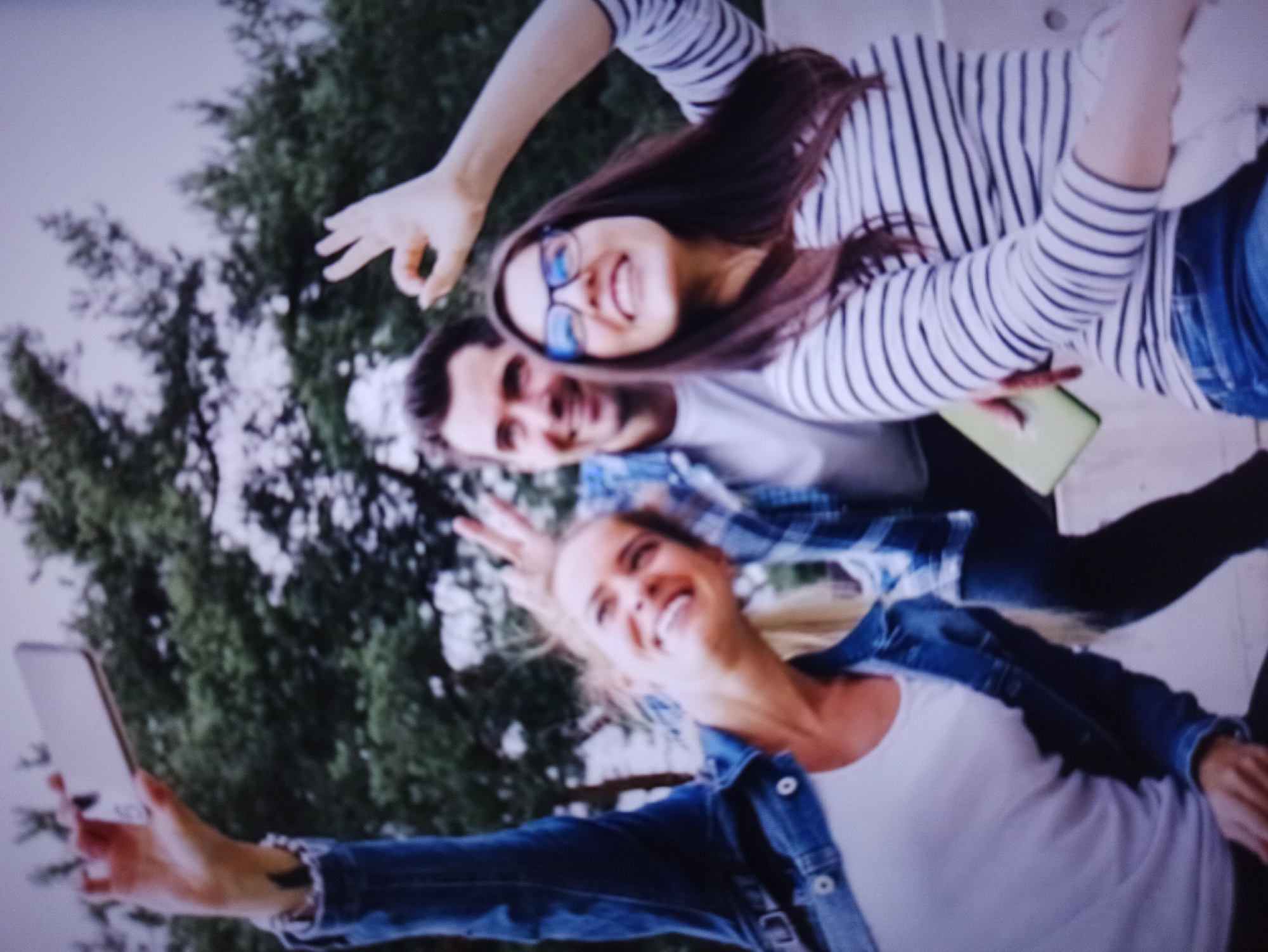 Three people are taking a selfie outdoors. The person on the left, a woman with blonde hair wearing a denim jacket over a white t-shirt, holds up a smartphone. To her right is a man with dark hair and a beard, wearing a blue and white checkered shirt over a white t-shirt. On the far right is a woman with long dark hair and glasses, wearing a striped long-sleeved shirt. She is making an "okay" gesture with her right hand. The man is also making an "okay" gesture with his right hand. They are all smiling. The background consists of trees and an overcast sky. The location is not identifiable as Balakliya, Ukraine.
