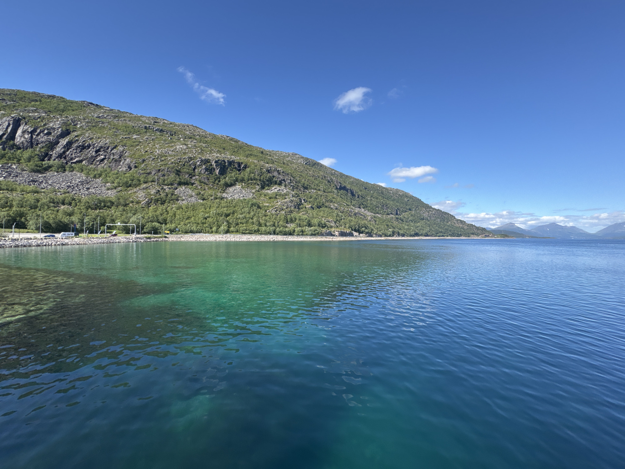 This media file is a landscape photograph taken in Lødingen, Norway, under a clear blue sky with a few wispy clouds. The scene depicts a body of water in the foreground, with a mountain range covered in greenery and rocky outcrops forming the backdrop. The water transitions from a shallow, turquoise hue near the shore to a deeper blue as it extends into the distance. Sunlight illuminates the water, revealing the rocky seabed in the shallower areas.
Along the shore, a road is visible, with several vehicles parked, including a white van and a dark-colored car. Trees and vegetation line the base of the mountain, creating a lush green band between the water and the rocky slopes. The mountain itself is characterized by steep, rugged terrain with scattered patches of vegetation.
In the far distance, other mountains can be seen, some capped with snow, suggesting a cool climate or higher altitude. The overall impression is one of natural beauty and tranquility, with a sense of open space and a bright, sunny day. The time of day appears to be mid-morning or early afternoon, given the strong sunlight and clear sky. There is no text visible in the image.