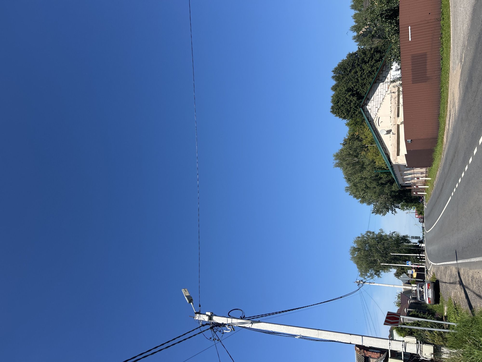 This is a low-angle, full shot of a paved road curving through a rural area under a clear blue sky in Kalitsino, Russia. A concrete utility pole with numerous wires and a street lamp is prominently positioned on the left side of the road. Adjacent to the pole, a small car is parked on the grassy shoulder. Further down the road, trees line both sides, and several houses with fences are visible. The road has white dashed lines marking the lanes. The overall environment suggests a sunny day in a village or suburban setting.