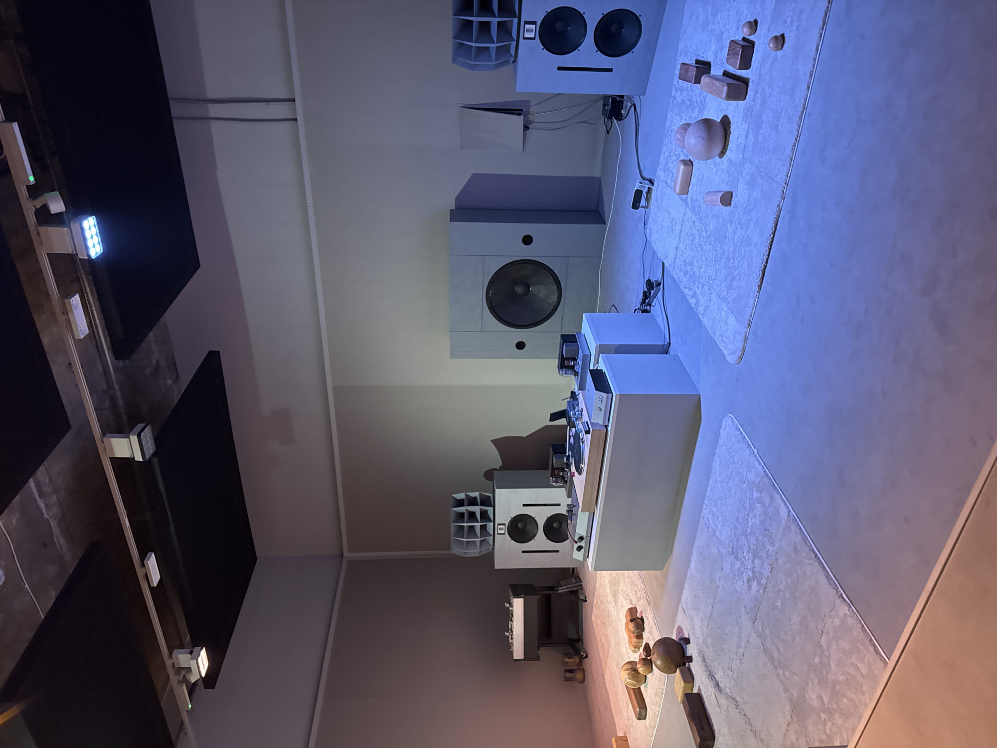 The image shows a recording studio or listening room, likely located in London, United Kingdom. The room is dominated by audio equipment. The walls are painted in neutral tones, and there are black acoustic panels hanging from the ceiling to control sound reflections.
Prominent features include large, high-end speakers, some paired with horn tweeters. Electronic components, such as amplifiers or preamps, are visible on white cabinets. A turntable and recording equipment are also present. The room has a cold color temperature, likely due to the lighting.
There are no people visible in the image.