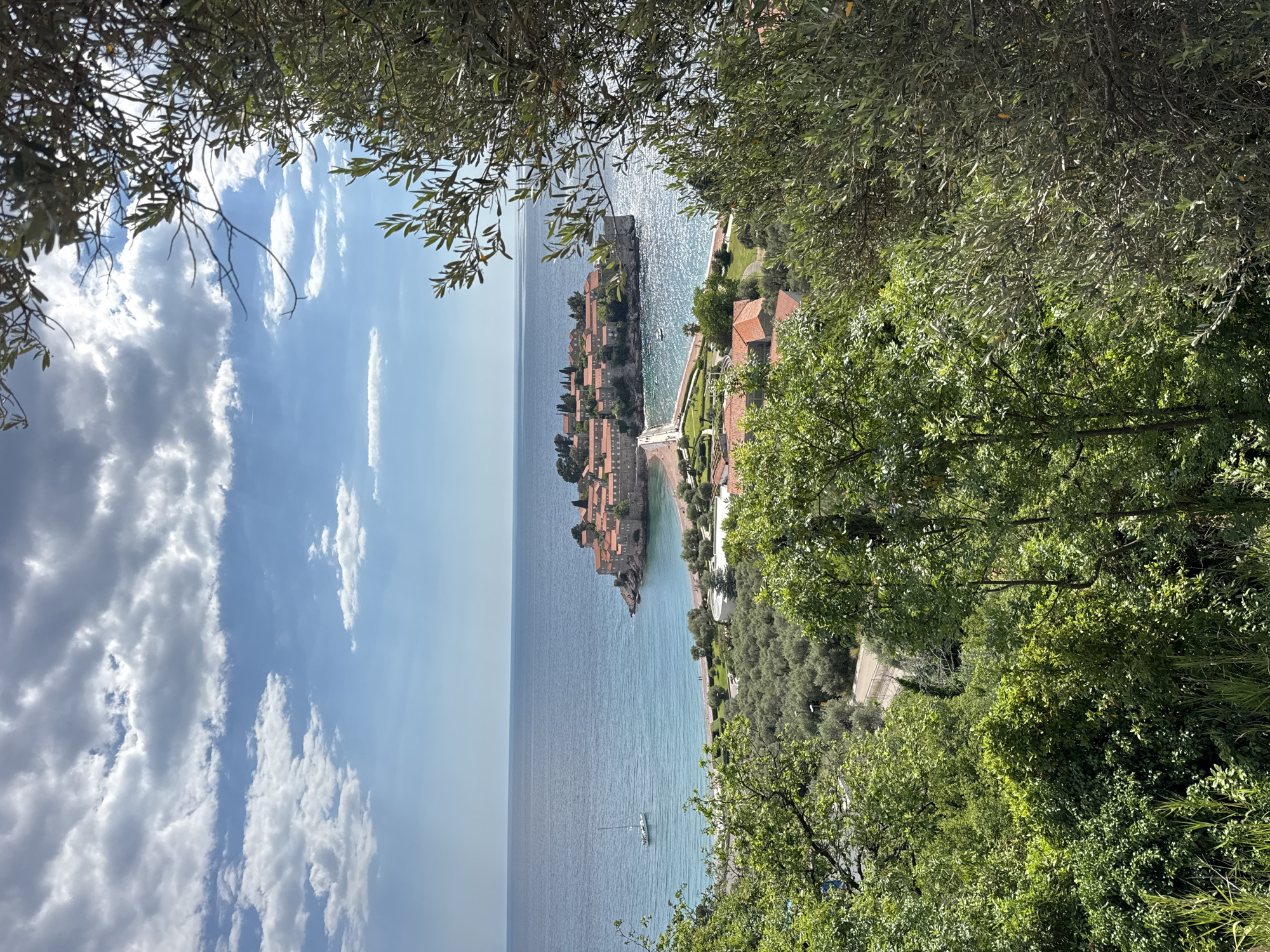 The image shows an elevated view of Sveti Stefan, Montenegro, framed by foliage in the foreground. The turquoise sea dominates the scene, with a sailboat visible. Sveti Stefan, a small islet connected to the mainland by a narrow isthmus, features buildings with terracotta roofs. A walkway connects the islet to the coast, lined with greenery. The sky is partly cloudy, with white clouds against a blue backdrop.