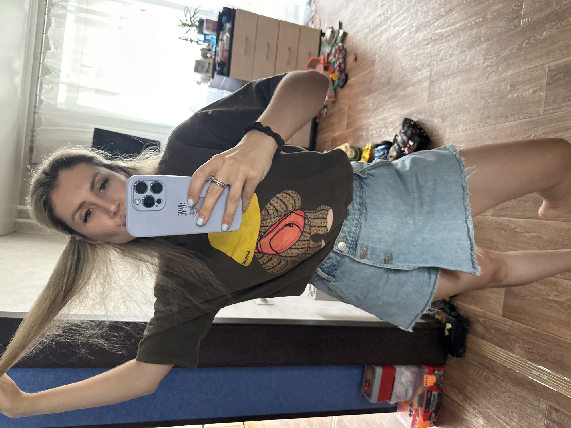 A woman is taking a selfie in a room. She has long blonde hair and is wearing a dark green t-shirt with a graphic design and a light blue denim skirt. She is holding a light purple phone in her right hand, which is reflecting in a mirror. The room has wooden floors and a blue rug. There is a dresser with drawers in the background, and some toys are visible on the floor. The lighting suggests it is daytime. The woman appears to be playful, puckering her lips for the photo. Visible text on the phone case reads "ВИИИТИЛ".