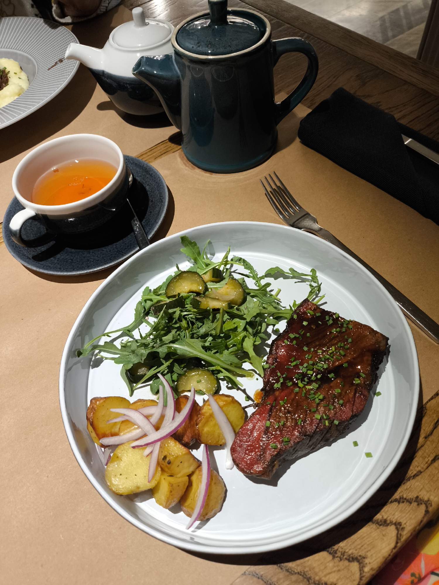 This image depicts a dining table scene in Izhevsk, Russia, featuring a prepared meal and tea service. The central focus is a white, oval-shaped plate positioned on a light brown paper or fabric table covering, which overlays a dark wooden table surface.
On the main plate, a dark reddish-brown steak, glistening with a sauce and garnished with chopped green chives, occupies the right side. To the left of the steak are several golden-brown roasted potato chunks, interspersed with thin slices of red onion. The upper left portion of the plate holds a green salad consisting primarily of arugula leaves mixed with sliced dill pickles.
To the left of the main plate, a dark grey ceramic teacup containing an amber-colored liquid (tea) sits on a matching saucer, with a small spoon resting within the saucer. Behind this teacup are two ceramic teapots: a white teapot with a dark grey base and lid, positioned slightly to the left, and a dark teal teapot with a matching lid, situated to the right.
A silver four-tined fork is placed to the right of the main plate. In the upper right corner of the table, a black folded cloth napkin conceals a partially visible silver knife. A portion of a white plate with a patterned rim, containing mashed potatoes topped with a dark substance, is visible in the upper left corner of the image, along with a blurred segment of a human hand or arm. The background shows blurred elements consistent with a restaurant interior, including wooden textures and a hint of a colorful, abstract pattern in the lower right corner.