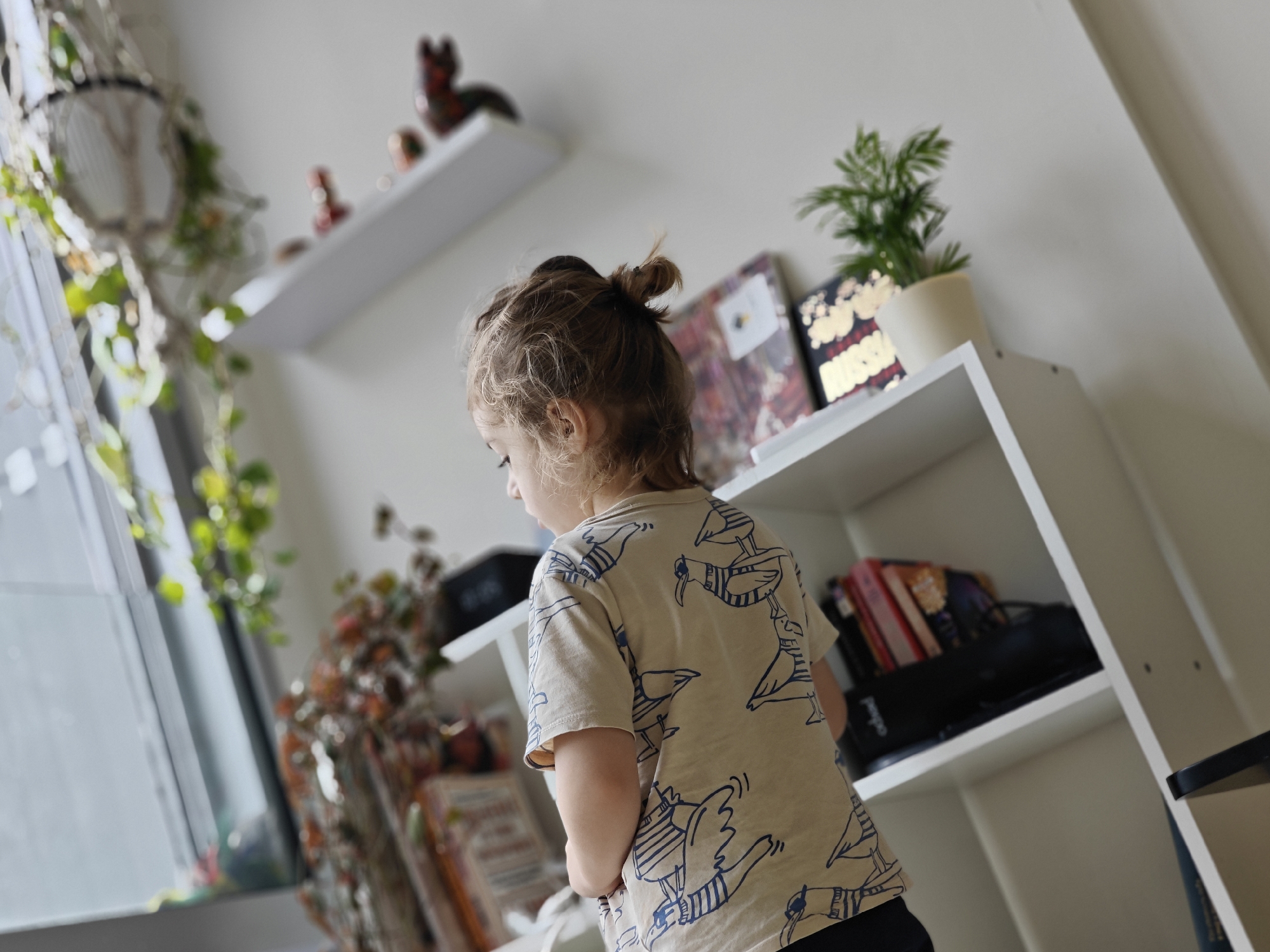 A young child, seen from behind, stands in front of a white bookshelf. The child has blonde hair styled in a bun and is wearing a beige t-shirt with a blue bird pattern. The bookshelf contains several items, including books, a small potted plant, and decorative figurines. To the left of the bookshelf, a window with a macrame plant hanger is visible. The room appears to be a home interior. The image does not contain any elements that would indicate its location in Mumbai, India.