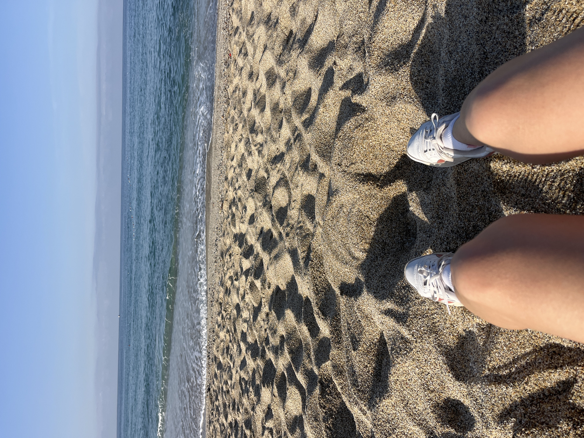 The image shows a view from the perspective of a person standing on a beach. The lower portion of the frame captures the person's legs and feet, wearing white sneakers and white socks. The beach consists of golden sand with contrasting shadows. Beyond the sandy foreground, the sea extends horizontally across the frame. The sky is visible in the upper portion of the image, appearing light blue. Based on the beach and sea, the image location is likely within the coastal city of Alanya, Turkey.