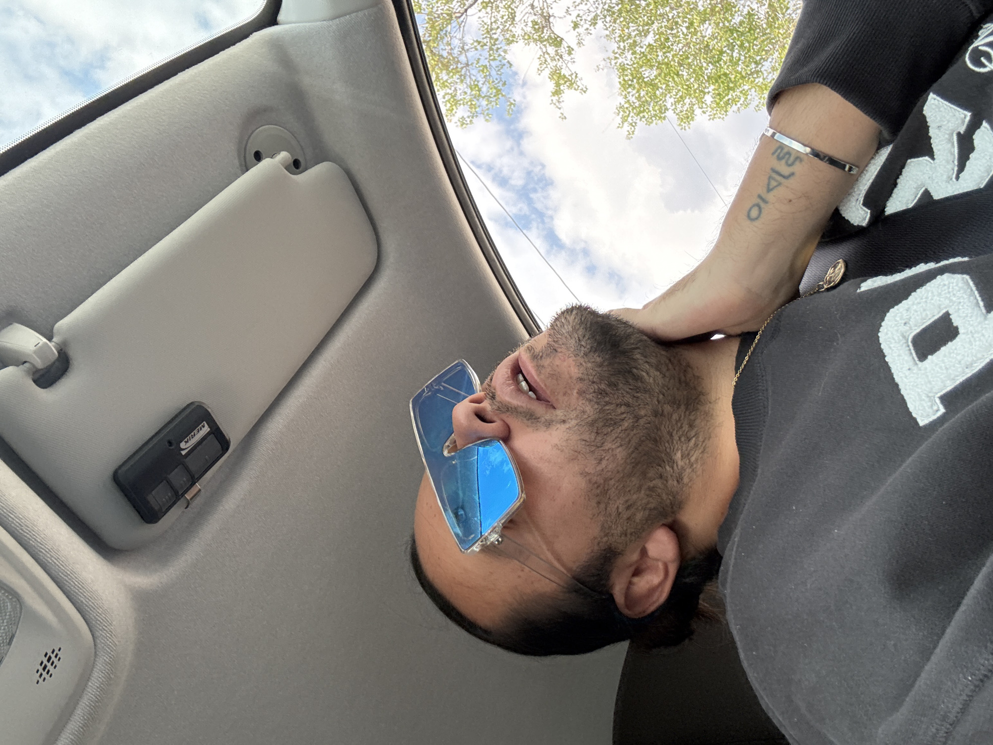 A man with a beard and blue sunglasses is positioned inside a car, looking up at the sky visible through the windshield. The car's interior, including the sun visor and roof, is visible. He is wearing a black sweatshirt, a silver bracelet, and a gold necklace. His left hand is placed on his neck. The scene appears to be outdoors with trees visible in the background. The specific location is not ascertainable.