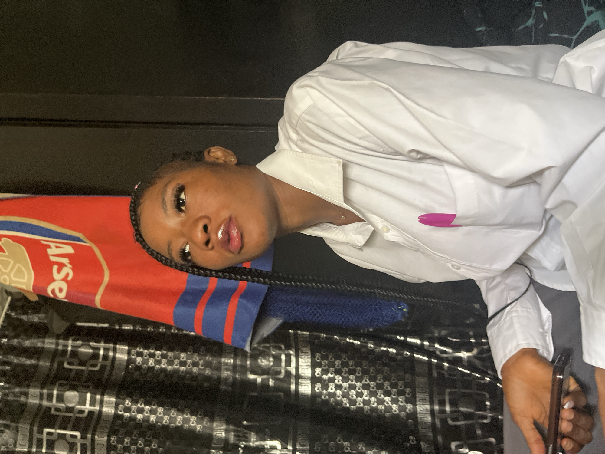 A young Black woman with braided hair is looking upwards and to the right, with her mouth slightly open. She is wearing a white button-down shirt with a pink object tucked into the breast pocket. She is holding a dark-colored smartphone in her left hand. Behind her, on the left, is a black patterned curtain or fabric. To her right is a red and blue striped banner that appears to be a sports team's flag, with the word "Arse" visible on it, likely referring to Arsenal Football Club. The background is dark, suggesting an indoor setting. The image was taken in the city of Jos, Nigeria.