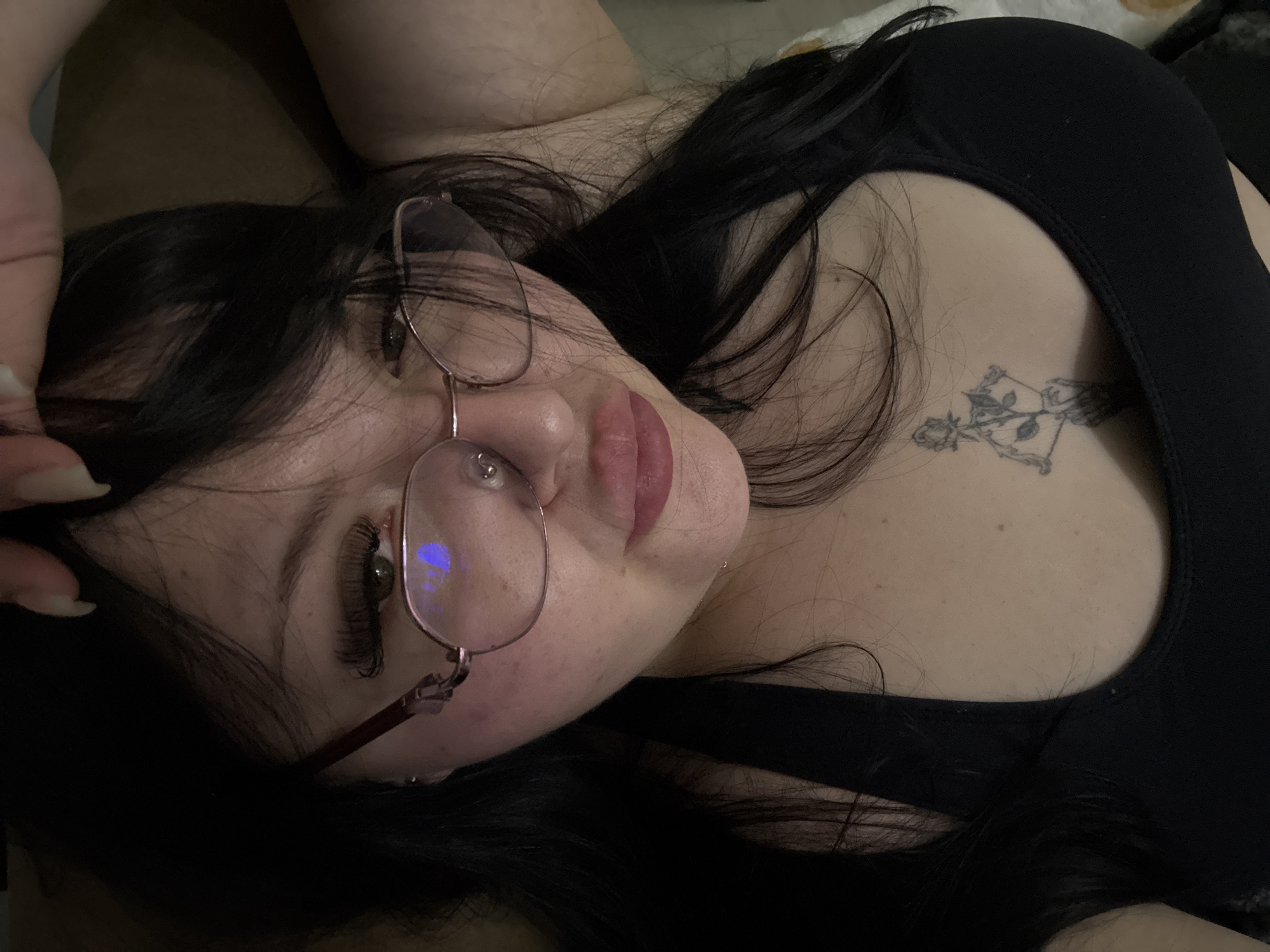 A woman is lying down, wearing glasses, a black tank top, and with a rose tattoo on her chest. The location is not discernible from the image, so it's impossible to determine if it's in Sofia, Bulgaria.