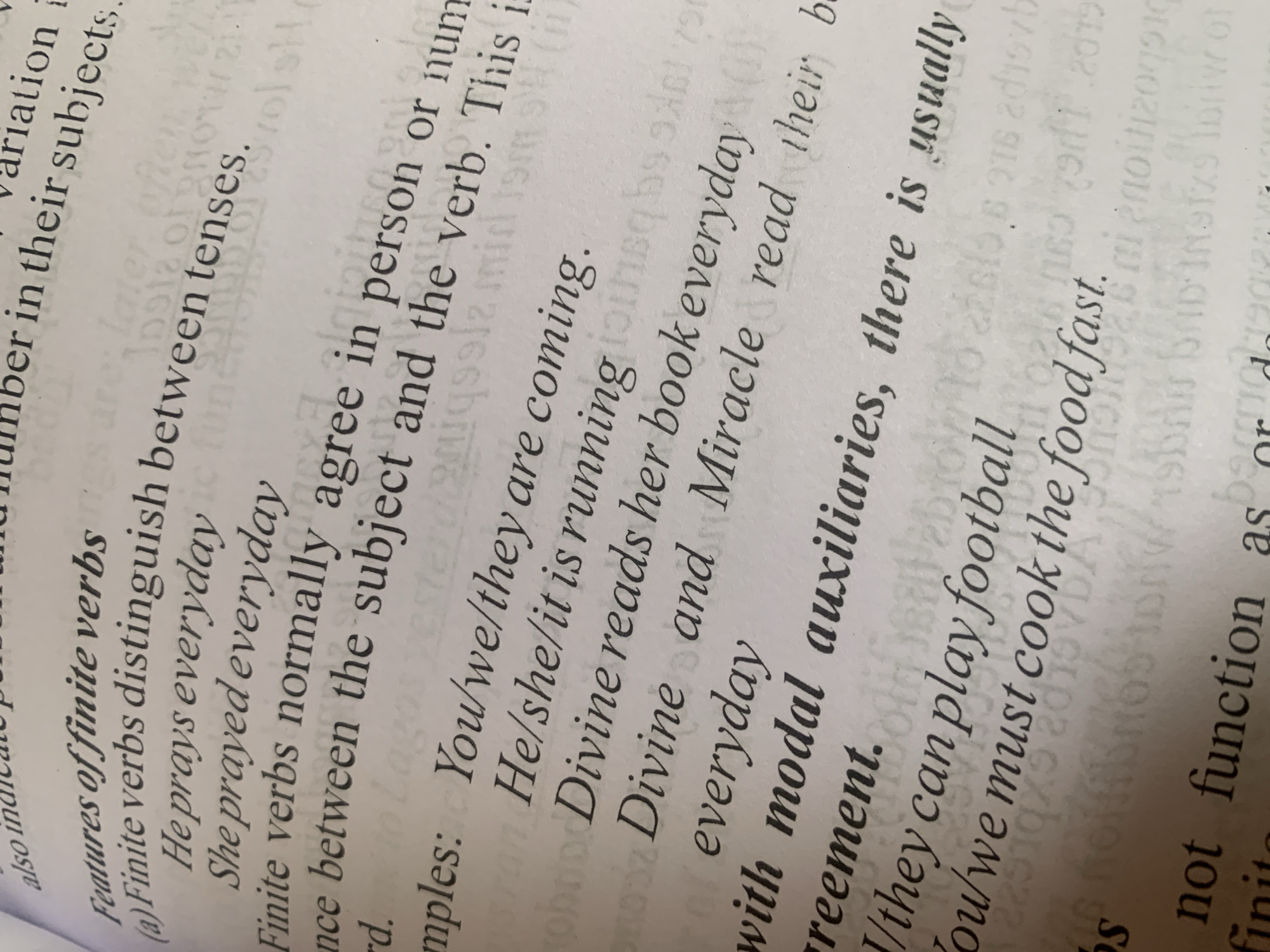 The image shows a close-up, angled view of an open book, focusing on text related to grammar. The text discusses "Features of finite verbs" and provides examples of sentences. It also touches on "modal auxiliaries" and their agreement. The lighting is uneven, with some areas brighter than others, and the page appears slightly creased. There are no people, animals, or discernible locations visible in the image. The background is indistinct, appearing as a blur of paper or possibly another page. The text discusses grammatical concepts like tenses, person, number, and agreement between subjects and verbs. There is no information about Jauro Yinu, Nigeria, present in the image.