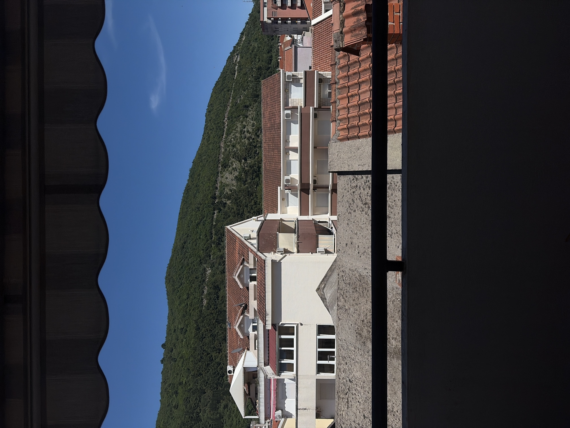 View from a building in Budva, Montenegro, including a section of dark roof, a mountain covered in dense green trees, a clear blue sky, and an apartment building with red tile roofs and white exterior walls. A section of wall and a metal bar are on the right.