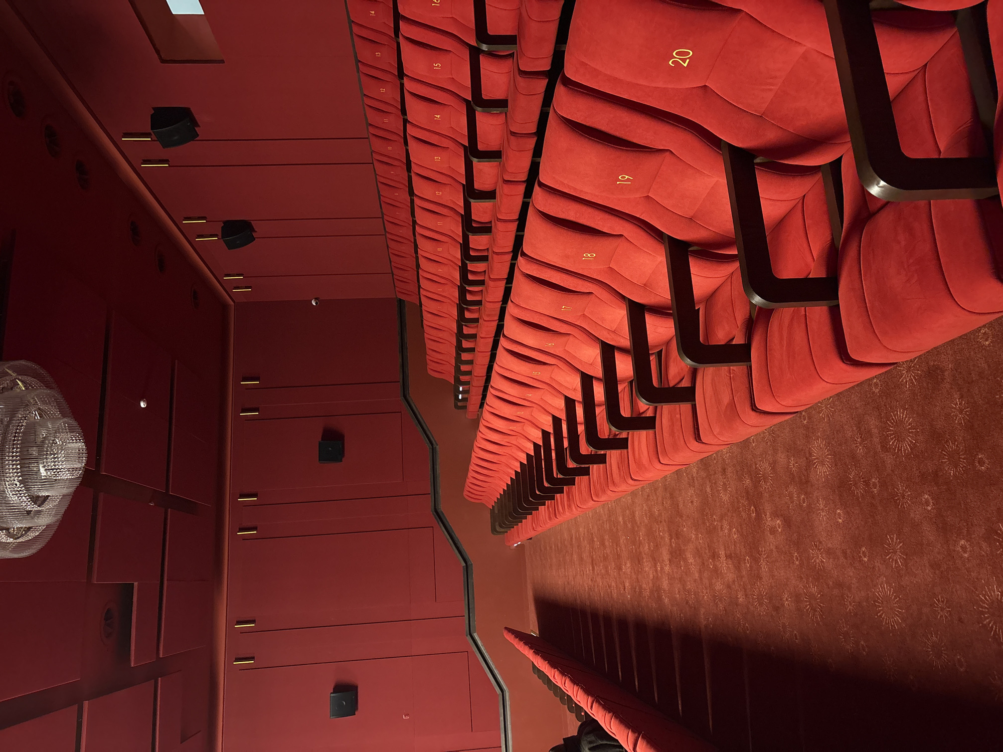 The image shows the interior of a theater hall, likely a cinema or performance venue. The main subject is the rows of red velvet seats, numbered in gold from 16 to 20. The seating is arranged in ascending rows, receding into the distance. The floor is covered with a patterned carpet in shades of red and brown. The walls and possible access doors visible to the left are painted in the same red hue. A disco ball is partially visible in the upper left corner of the frame. Based on the information provided, the location is Minsk, Belarus.