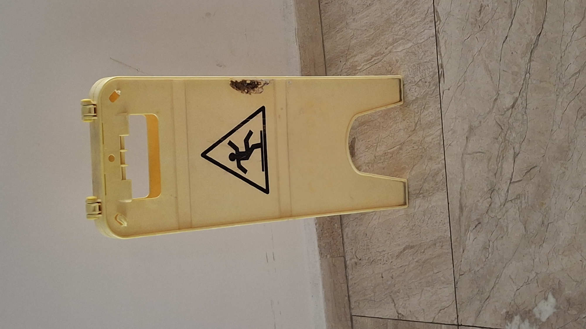 A yellow, plastic wet floor sign stands on a beige tiled floor in Nesebar, Bulgaria. The sign features a black symbol of a person slipping on a wet surface within a black triangle. The sign has a cutout handle at the top and two leg-like bases. There is a small, damaged area on the right side of the sign. The background consists of a white wall with a light beige baseboard.