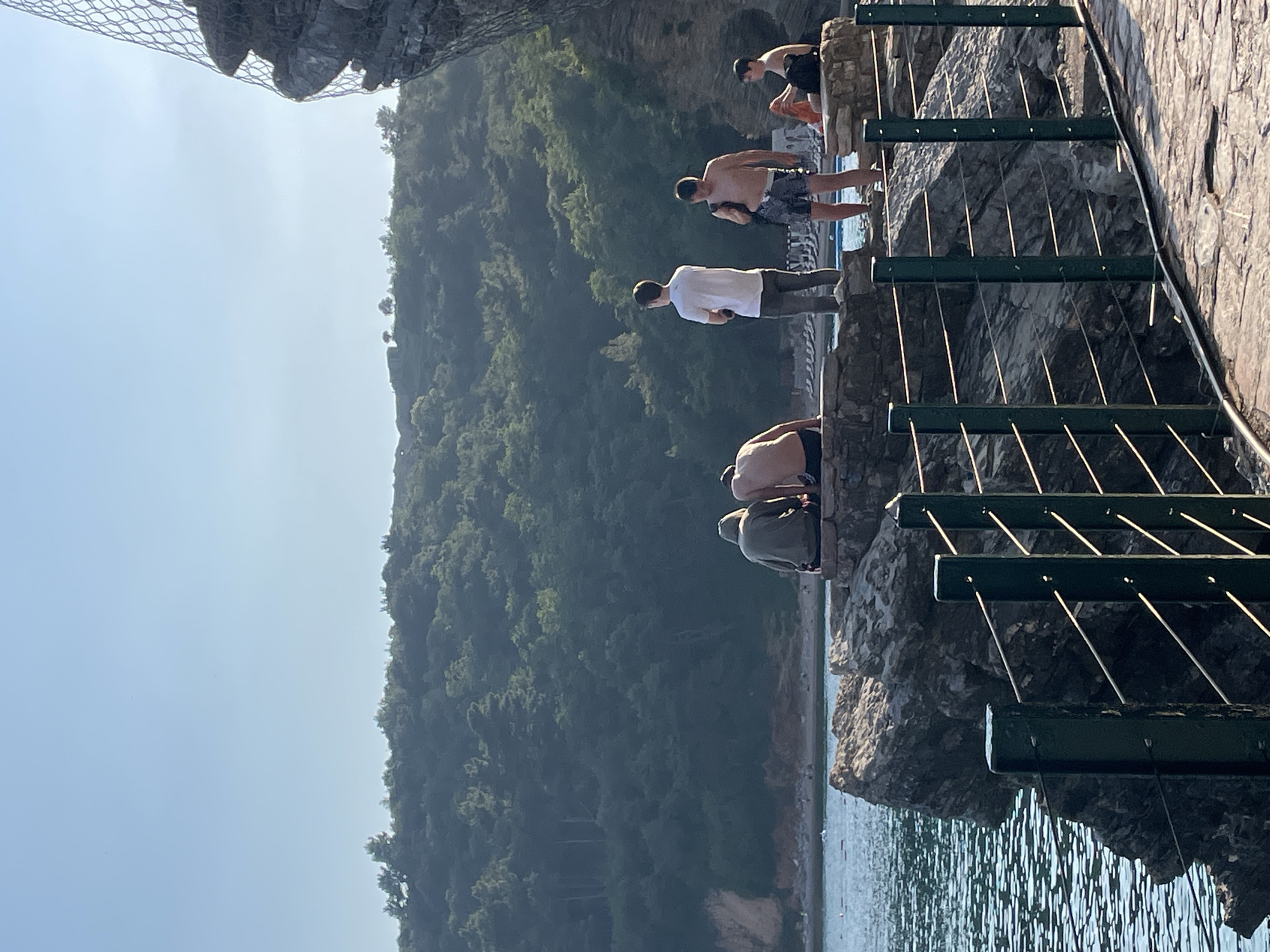 A sunny, daytime shot taken along a coastal path in Budva, Montenegro, features a group of six people standing near a rock face above the Adriatic Sea. The path appears to be constructed of stone, bordered by a low wall with green metal railings. From left to right, the subjects are described as follows:
* Two individuals are near the wall, possibly preparing to jump or swim in the water below.
* A person in a white shirt and darker-colored pants faces the water.
* A person is wearing swim trunks.
* A person wearing swim trunks is standing on the path.
* One person standing by the wall is wearing dark clothing.
Behind the people, a steep, green hillside covered in trees rises sharply. The sky is light blue with a slight haze. The angle of the photograph is slightly tilted. The net is visible on the upper left side.