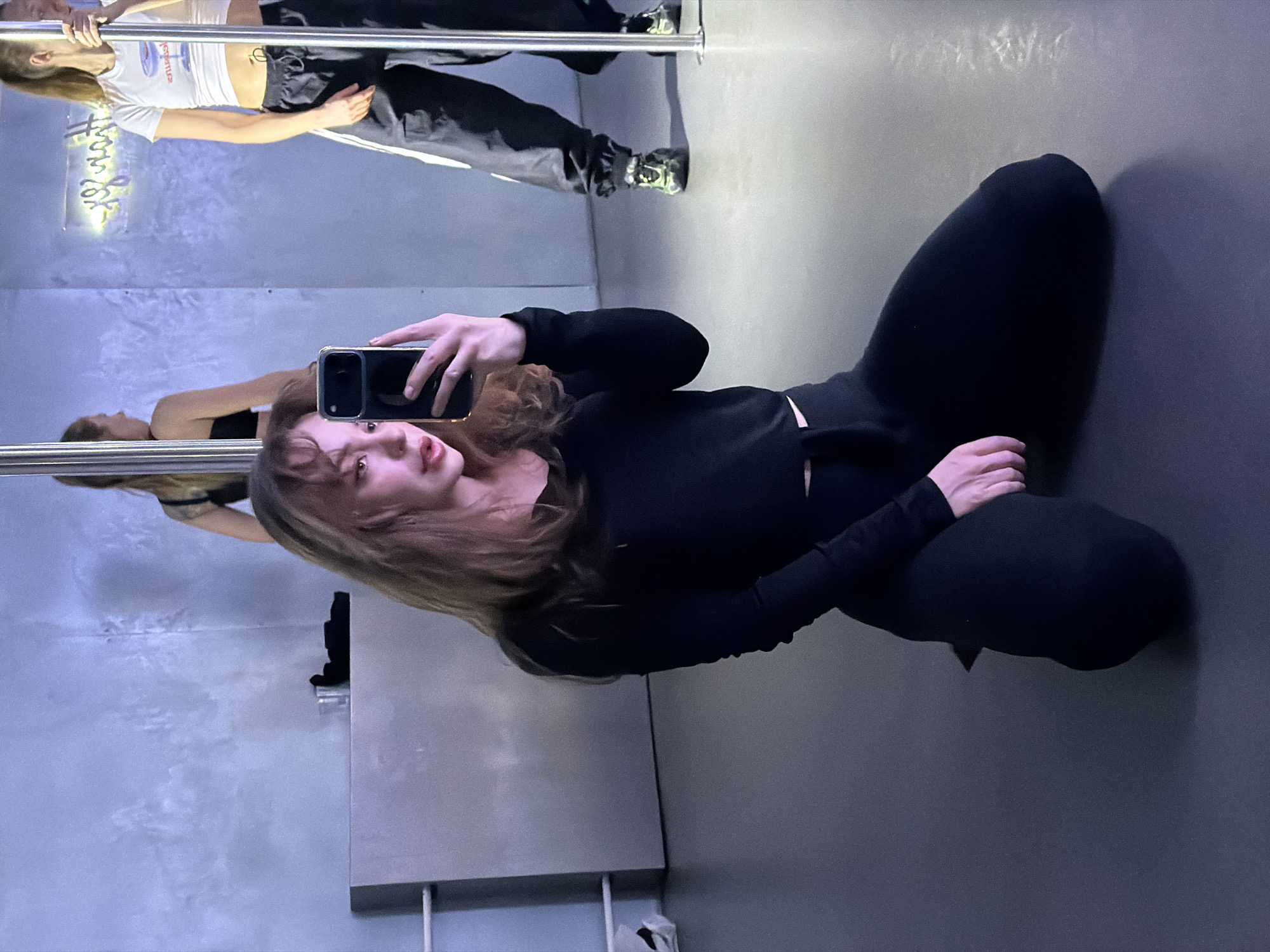 This image depicts an indoor scene within a pole dance studio located in Moscow, Russia.
In the foreground, a young adult female with light brown hair and bangs is kneeling on a dark gray floor. She wears a black long-sleeved crop top and black fitted trousers. Her right hand holds a dark-colored smartphone with a light bezel, positioned to take a selfie. Her lips are slightly parted.
In the background, a large mirror serves as the back wall. Reflected in this mirror, to the left, another young adult female is visible from the upper back, wearing a black strapless top or sports bra, standing near a chrome vertical pole. To the right, a third young adult female stands holding a chrome vertical pole with her left hand. She wears a white cropped t-shirt with a colorful graphic, black baggy cargo pants with white side stripes, and neon green and black athletic shoes. Her light brown hair appears tied back.
A low, silver-colored counter runs along the base of the mirrored wall. On this counter, a clear glass and a folded black item are visible. Above the counter, an inverted neon sign reflected in the mirror displays the English words "POLE DANCE." The room is lit with a cool, blueish-purple ambient light.