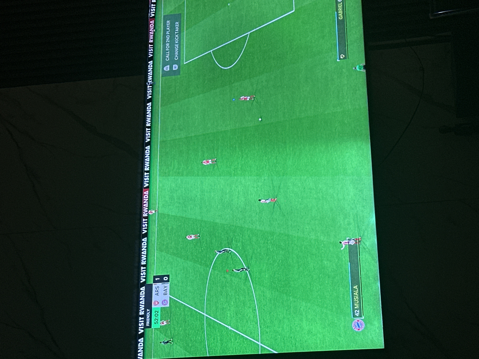 The image shows a screenshot of a soccer video game being played on a television. The game features a match between Arsenal and Bayern Munich, with Arsenal leading 1-0 at 52:02 into the game. Players from both teams are visible on the green field. The background of the screen displays "VISIT RWANDA" branding repeatedly. On the lower right side of the screen, there are game control prompts. In the lower left corner, a player's name, "42 MUSIALA", is displayed along with the Bayern Munich logo. On the lower right, a player's name, "GABRIEL", is displayed. This scene is not located in Gwarinpa, Nigeria; it is a virtual environment within a video game.
