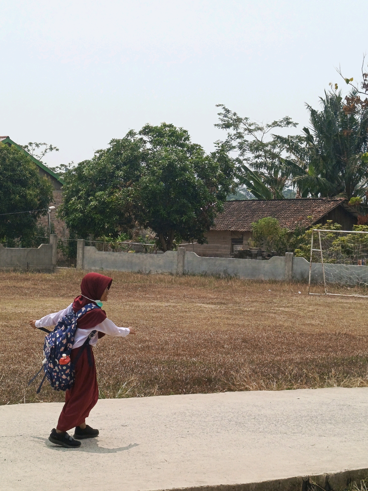 A young girl wearing a maroon hijab, white shirt, and maroon pants, with a blue backpack adorned with cloud and moon patterns, is walking on a concrete path. She is carrying what appears to be a water bottle in a side pocket of her backpack. The girl is facing away from the camera, looking towards the right. The background shows a dry, brown grassy field, a concrete wall, and a wooden house with a tiled roof. Behind the house, there are trees, including palm trees. A white goalpost for a soccer field is visible to the right of the house. The sky is overcast and pale. The scene is set in a rural or semi-rural area, consistent with locations like Megang Sakti V, Indonesia.